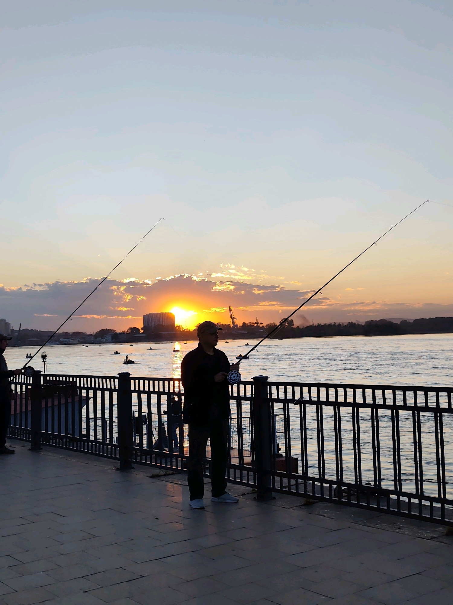 The image depicts a waterfront scene at sunset. A man, silhouetted by the setting sun, stands on a paved walkway, holding a fishing rod. He is wearing a cap, a dark jacket, and dark pants. A black metal fence separates the walkway from the water. In the background, the sun is setting behind a cluster of buildings and a crane, casting a warm glow across the sky and clouds. The water is calm, with several small boats scattered across it. The overall atmosphere is serene, with a sense of twilight tranquility.