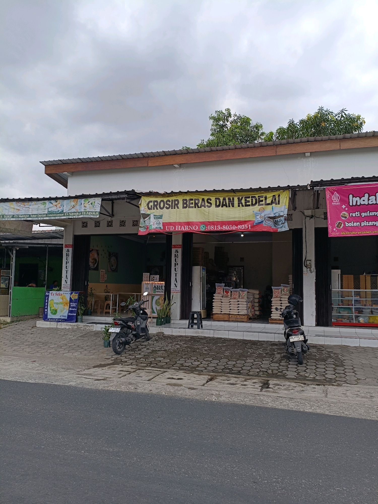 The image captures a storefront in Maguwoharjo, Indonesia, on an overcast day. The building appears to be a commercial establishment with a white facade and a corrugated metal roof. Two motorcycles are parked in front of the building on a paved walkway.
On the left side of the building, there is an open area with green walls and a sign advertising various iced beverages, including "Es Teler," "Es Mango," and "Es Alpukat." A banner at the top of this section lists more beverage options and the word "SRUPUTAN."
The central part of the building features a large storefront with metal roll-up doors, which are partially open. Above the entrance, a yellow banner displays "GROSIR BERAS DAN KEDELAI" (Wholesale Rice and Soybeans) and "UD HARNO" along with a phone number. Inside, stacks of rice sacks are visible.
To the right, another section of the building has a pink banner with the text "Indah." Below this banner, a menu lists "roti gulung" (rolled bread) and "bolen pisang" (banana bolen), indicating it's a food-related business. A display shelf with products is visible inside this section.
The foreground shows a paved area in front of the storefront, transitioning to a grey asphalt road. The environment is typical of a commercial street.