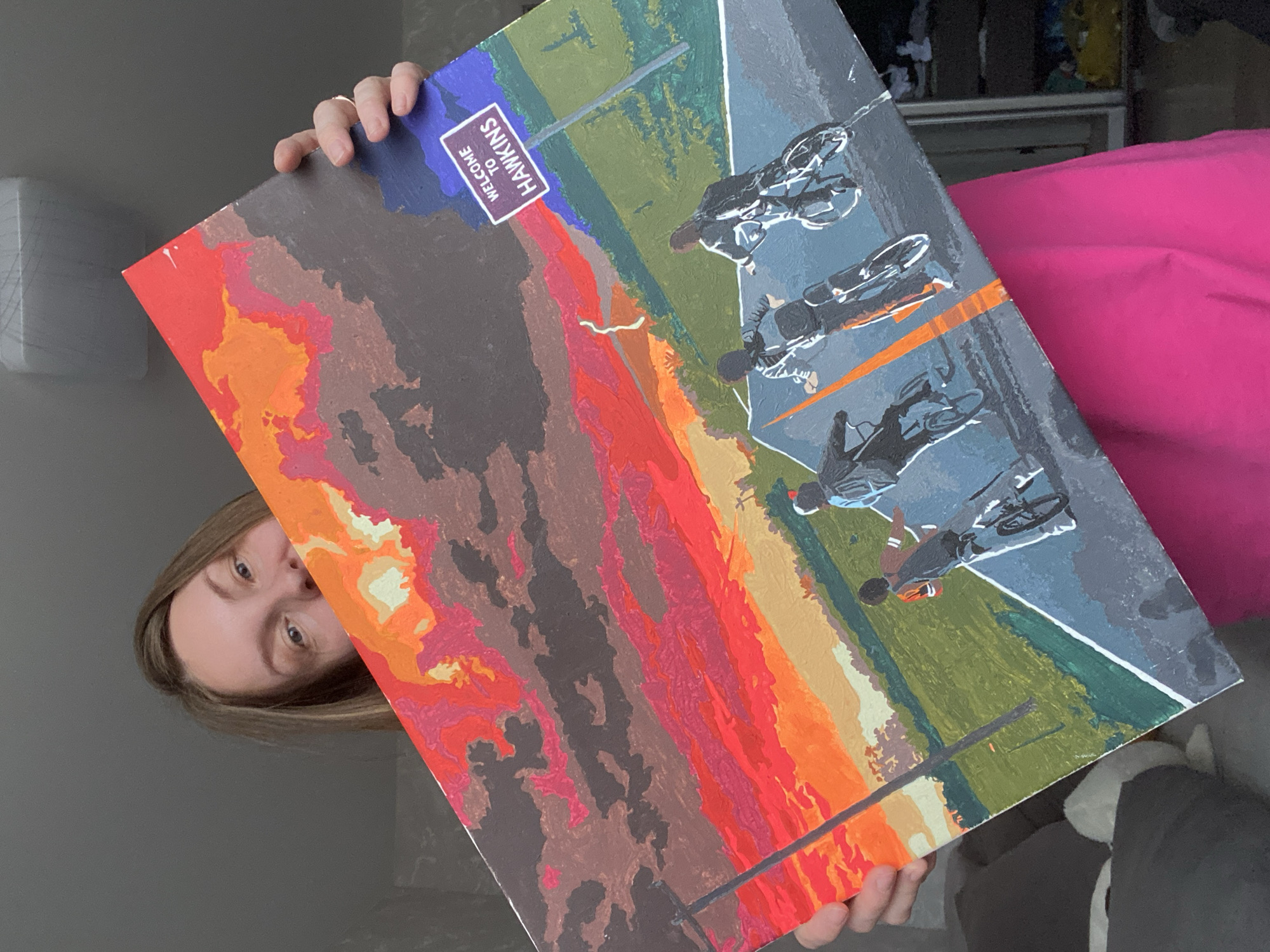 A person is holding a painting of a scene reminiscent of the Netflix series "Stranger Things." The painting depicts a "Welcome to Hawkins" sign, a group of people on bicycles riding on a road, and a fiery landscape in the background. The painting is being held indoors. The person's face and part of their body are also visible. The background includes a wall, a white object, and part of a closet or wardrobe.