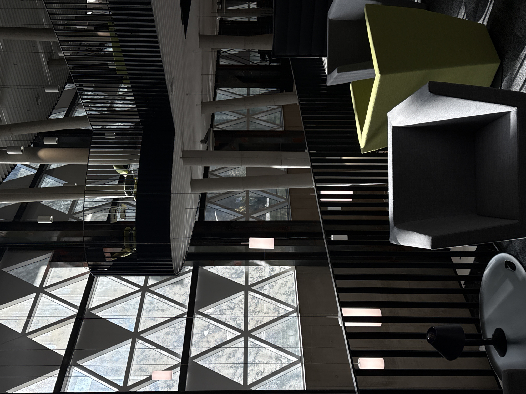 The image depicts the interior of a modern, multi-level building located in Esch-sur-Alzette, Luxembourg. The scene is characterized by striking contemporary architecture, featuring a prominent, large-scale window system composed of numerous interconnected triangular glass panels that form a geometric pattern on the main wall. These windows allow substantial natural light into the space, creating strong contrasts between brightly illuminated areas and deep shadows, particularly on the dark floor covering.
The interior is multi-tiered, with at least two visible levels connected by an open space featuring black vertical-bar balustrades that delineate walkways and an upper-level balcony. Several large, light-colored, cylindrical support columns, some appearing to be angled, extend from the floor to the ceiling, providing structural support. The ceiling is flat, light-toned, and features multiple cylindrical pendant lights suspended at various lengths, some of which are illuminated.
On the foreground of the lower level, a seating arrangement includes modern, angular upholstered armchairs. One dark grey armchair is prominent, situated next to a round, metallic-finished low table with a raised edge, on which a small, black conical desk lamp is placed. Directly behind this, an olive green upholstered armchair is visible, followed by a partially obscured, tall, dark upholstered high-backed seating unit. Through the black balustrades, additional seating units, including more olive green chairs, can be seen on the upper level. No individuals are present in the image, and no actions or interactions are depicted, rendering the scene static and unoccupied.