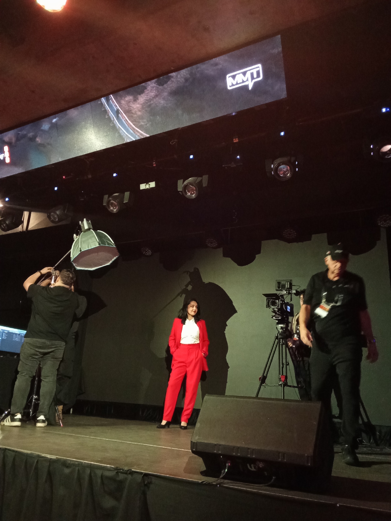 The image depicts a stage setup with a large screen displaying the "MMT" logo. In the foreground, a woman in a vibrant red suit stands with her hands clasped behind her back, looking to her right. To her left, a person in a black t-shirt and grey pants is adjusting a studio light with a large diffuser. To the right of the woman, a videographer is operating a camera mounted on a tripod. Another person, wearing a black t-shirt, black cap, and black pants, walks across the stage from right to left, partially obscuring the videographer. The stage is equipped with several stage lights and a large speaker in the foreground. The background appears to be a dark curtain. The scene suggests a professional event or performance, possibly a presentation or filming. The image was taken in Santiago, Chile.