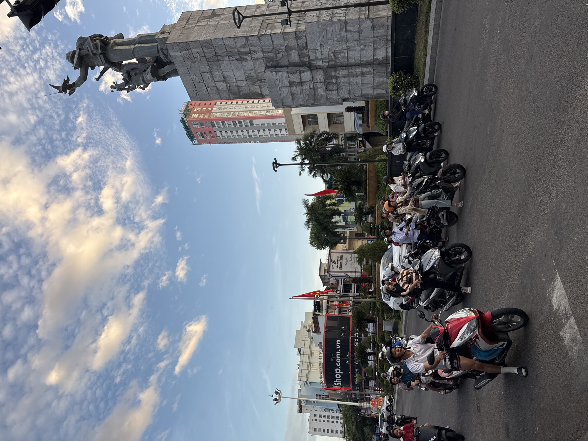 The image depicts a busy street scene in Nha Trang, Vietnam, at dusk. The foreground shows a traffic jam of motorbikes, with riders and passengers wearing helmets and casual clothing. A prominent statue, possibly a war memorial, stands tall on the right side of the frame, silhouetted against the twilight sky. Behind the statue, a modern, multi-story building with a red and white facade rises high into the sky. In the mid-ground, commercial buildings with signage, including "Shop.com.vn", are visible. The sky is a mixture of blue and light orange clouds, indicating the setting sun. Palm trees and other greenery are scattered throughout the urban landscape. The overall impression is of a vibrant, bustling city at the end of the day.