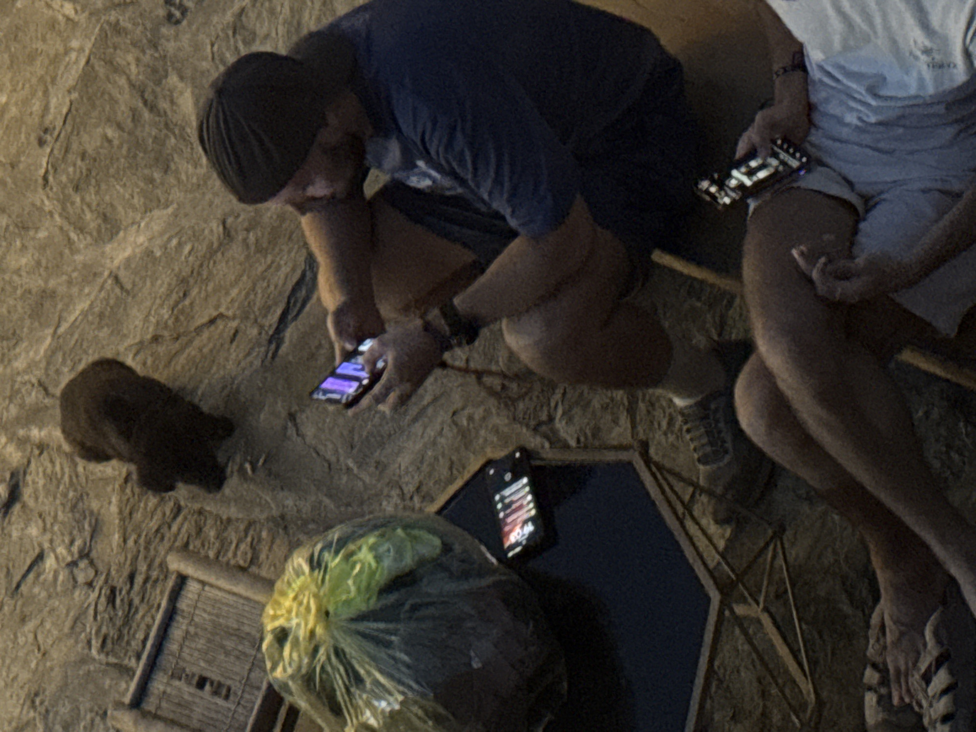 Two men are seated on a stone surface in what appears to be an outdoor setting, possibly in Bắc Hà, Vietnam. One man, wearing a dark t-shirt, shorts, and a dark cap, is crouched and looking at a smartphone in his hands. The other man, wearing a light-colored t-shirt and shorts, sits with a smartphone in his hand, glancing downward. There is a black object, possibly a dog, near the first man. A plastic bag containing leafy contents is present near a small black table with a phone on top. The overall environment appears to be dimly lit. The ground is uneven, possibly cobblestone or natural stone.