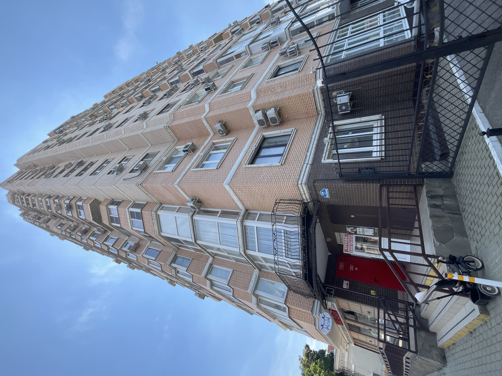 The image depicts a tall, multi-story brick apartment building under a bright blue sky with scattered clouds. The building has many windows, balconies, and air conditioning units visible on its facade. The perspective is from a low angle, looking up at the building, emphasizing its height.
At the base of the building, there are storefronts with signage. One sign appears to be for a dental clinic, featuring stylized teeth. Another sign indicates "АРЕНДА" (rent) with a phone number. A red door is visible, and next to it, a dark brown door. A metal fence with a decorative pattern encloses a section of the building's lower level, and a black metal gate is partially open.
In the foreground, on the sidewalk, a child's tricycle is parked near a set of stairs leading up to the building's entrance. The stairs are lined with railings. The sidewalk is paved with light-colored rectangular stones.
The location is identified as Krasnodar, Russia, based on the street name visible on a sign: "64 Ленина" (64 Lenin Street).