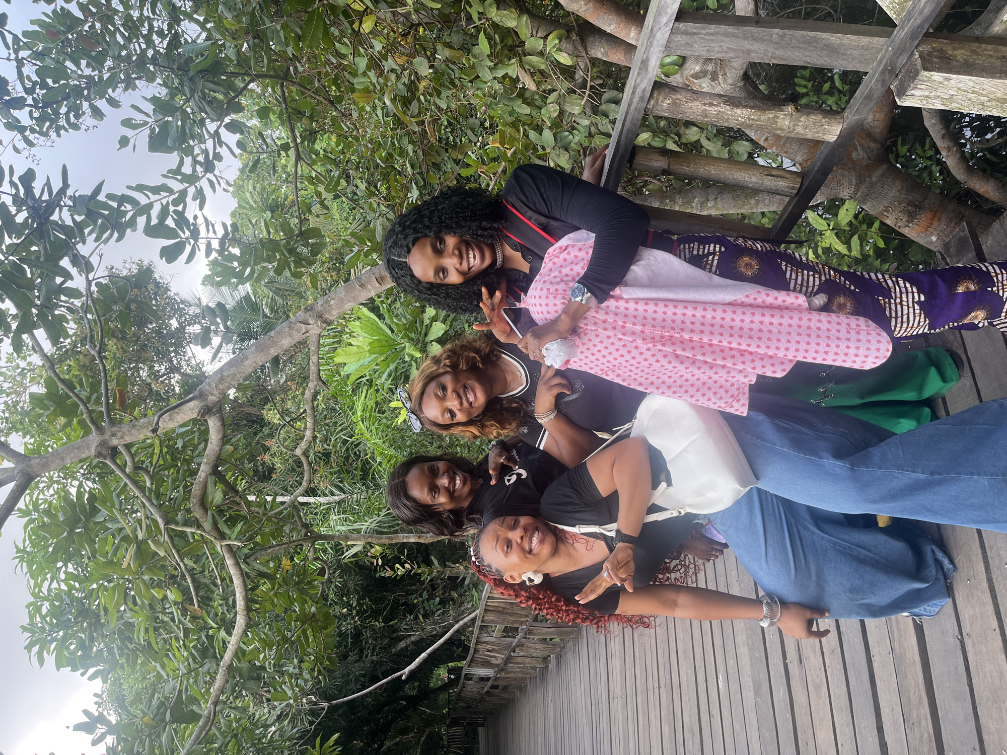 Four Black women are posing for a photo on a wooden boardwalk in a lush, green, natural environment. The boardwalk appears to be elevated, with wooden railings on the right side of the image. Dense foliage, including trees and various plants, surrounds the boardwalk. The sky is overcast.
From left to right:
The first woman on the left has bright red curly hair and is wearing large white hoop earrings, a black t-shirt, and very wide-leg light blue denim pants. She is smiling and has her left hand raised with two fingers extended in a peace sign, and her right hand is on her hip. She has a white purse slung over her shoulder.
The second woman from the left is wearing a black t-shirt and smiling broadly. She is pointing with her right index finger towards the viewer.
The third woman from the left is wearing sunglasses perched on her head, a black t-shirt with white lettering, and a necklace. She is smiling and has her right arm around the woman next to her.
The fourth woman on the far right has curly black hair, is wearing a black top with red trim, a pearl necklace, and a silver wristwatch. She is smiling and holding a pink and white polka-dot fabric, possibly a scarf or garment, in her arms. Her right hand is raised, and she appears to be waving or gesturing. She is wearing dark pants with a vibrant, geometric pattern.
The location is identified as Lekki, Nigeria, suggesting a tropical or subtropical climate given the dense vegetation. The boardwalk and surrounding natural environment suggest a park, nature reserve, or possibly a tourist attraction in the Lekki area.