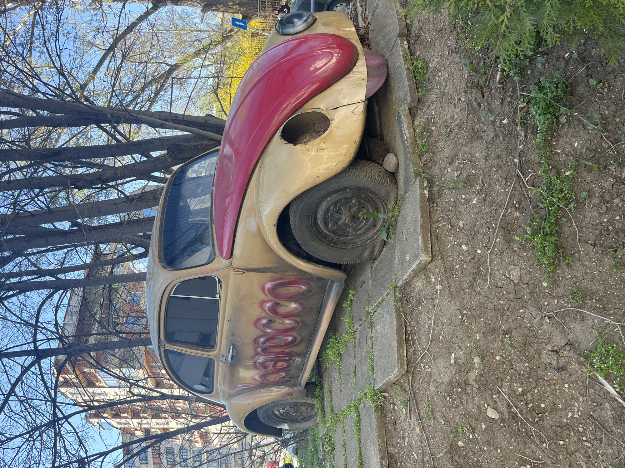 A partially stripped and graffiti-covered Volkswagen Beetle is parked in a small dirt area adjacent to a sidewalk in Sofia, Bulgaria. The car's front section is painted red and tan, with a large hole visible where the right headlight should be. A red graffiti design is seen along the side of the car. The car is wedged next to a concrete sidewalk lined with small plants. Bare trees are overhead, and a building is visible in the background. The time of day appears to be daytime under partly cloudy conditions. No people are in view, and no activities are taking place. The environment is urban and slightly neglected.