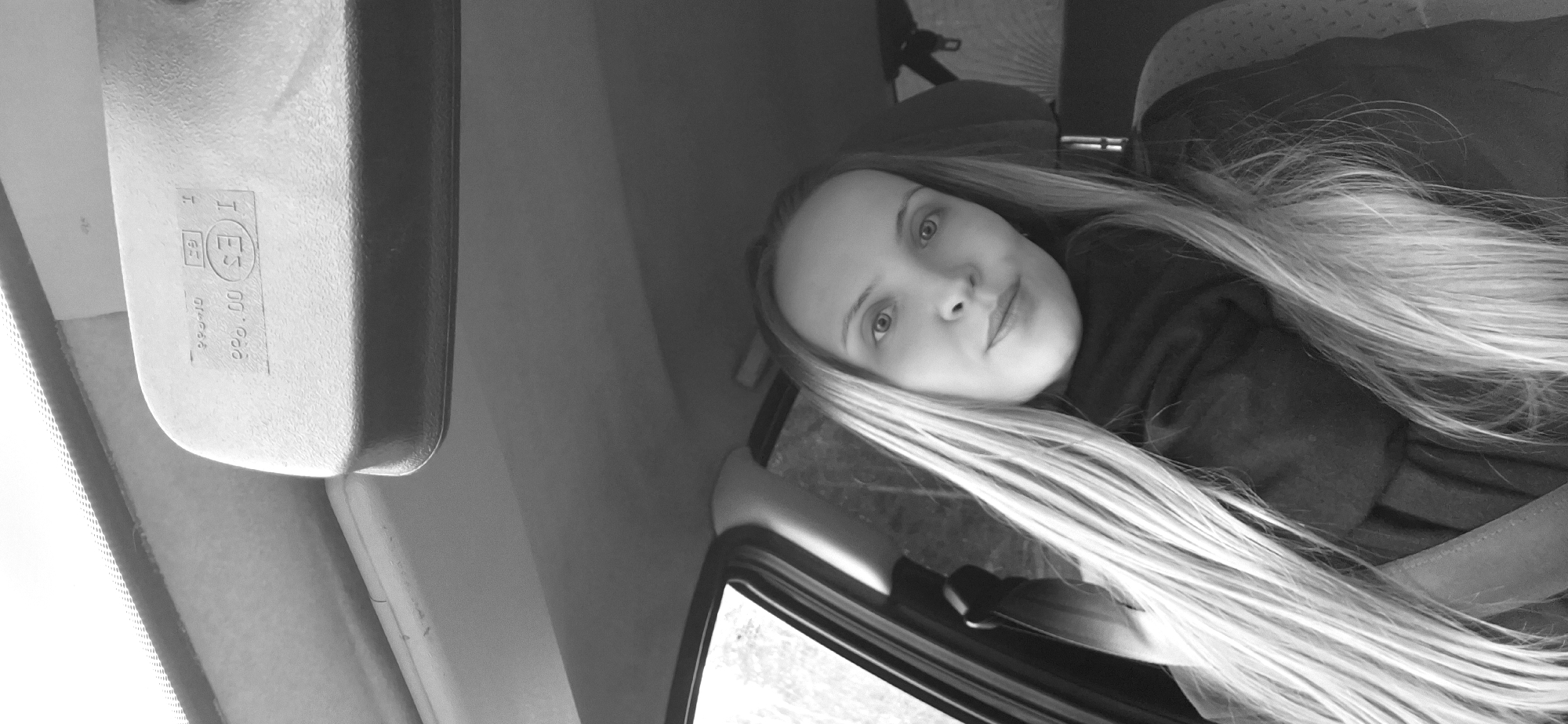 A monochrome, horizontally oriented image depicts a young woman inside a vehicle, likely a car. She has long, light-colored hair that flows over her left shoulder and arm, and is looking directly at the camera with a neutral to slightly pleasant expression. Her eyes are light-colored.
The shot is taken from within the vehicle, possibly a selfie. A rearview mirror is prominently visible in the upper left of the frame, showing various markings. Part of a vehicle window is seen on the right, through which some blurry outdoor scenery is visible, suggesting it is daytime. The interior of the car is otherwise simple, with parts of a seat or headrest discernible in the background.
Visible text on the rearview mirror includes: "I (ES)" within a circle, "00.000", "01-046", "GF" within a rectangle, and a solitary "H".
The context provided indicates the photo was taken in Chaikovsky, Russia.