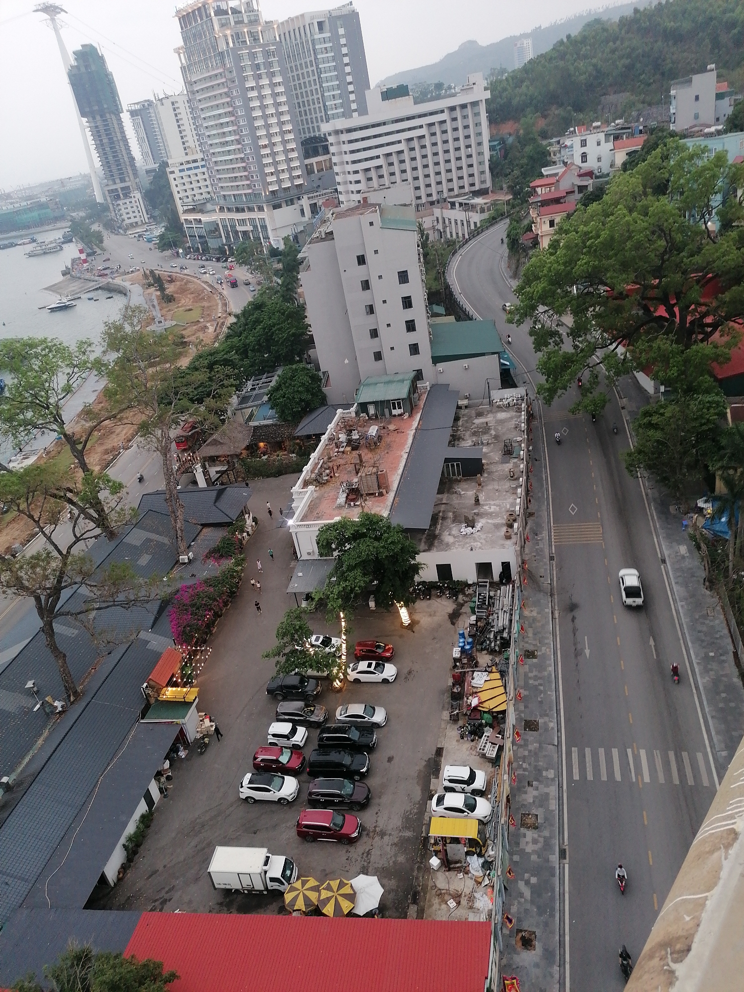 The image shows an aerial view of a section of Bai Chay Ward, Vietnam. Buildings ranging from modern skyscrapers to low-rise commercial structures define the urban landscape. A coastal road parallels the shoreline where several boats are docked. Inland, a winding road leads past densely packed buildings and verdant trees. A parking lot containing a mix of sedans, SUVs, and a small truck is visible, adjacent to a cluster of commercial buildings featuring outdoor seating and decorative lighting. The street below shows light vehicular traffic, including cars and motorbikes, and pedestrian activity. The sky appears overcast.