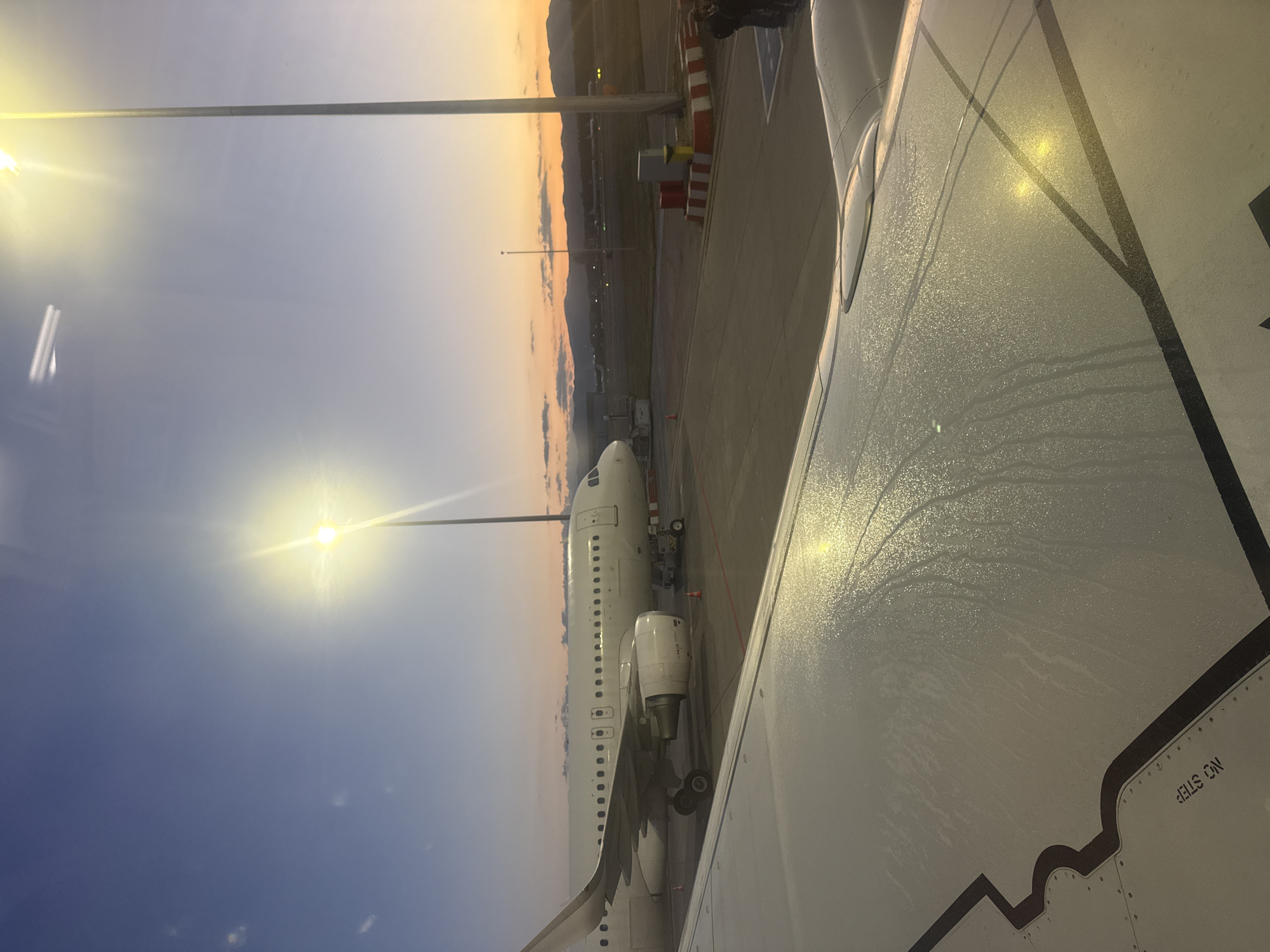 The image depicts an airplane wing in the foreground, covered in a pattern of water droplets, suggesting a recent rain or condensation. In the background, a white airplane is parked on the tarmac, with its engines and landing gear visible. The scene is set at dusk or dawn, with a colorful sky transitioning from blue to orange and yellow hues. Several light poles illuminate the tarmac, casting a warm glow. Mountains can be seen in the distance, silhouetted against the twilight sky. The overall impression is one of a peaceful airport scene during the transitional hours of the day.