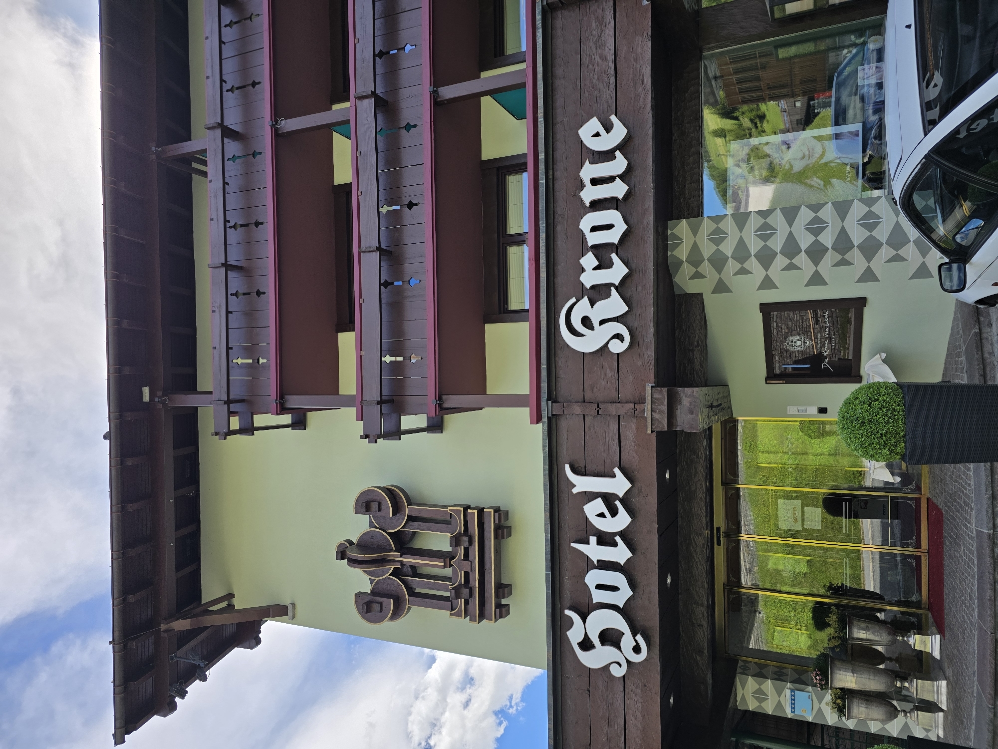 A hotel named "Sotel Krone" is depicted in Lech, Austria. The hotel has a light green facade and dark brown wooden balconies with intricate designs and burgundy trim. Above the entrance, "Sotel" and "Krone" are displayed in large, white, Gothic-style lettering against a dark wooden beam. A distinctive brown and gold emblem is mounted on the facade above the hotel name. The entrance features automatic glass doors with gold trim, flanked by large planters and urns. To the right of the entrance, a large window displays a black and white image of a woman and a car is parked in front. The surrounding environment is hilly and green, suggesting a mountainous landscape, consistent with the location in Lech. The sky is partly cloudy.