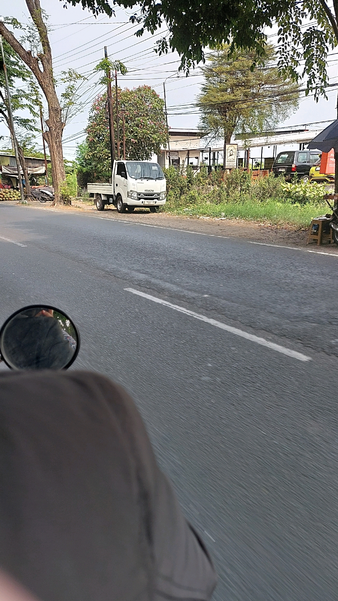 The image shows a white Isuzu truck parked on the side of a road in Ketimang, Indonesia. The truck is facing the viewer and is positioned on a dirt shoulder next to a paved road. In the background, there are several trees, some with green foliage and one with pink flowers. A building with a white facade and a covered outdoor seating area is visible behind the trees. To the left of the truck, there are some coconuts and a motorcycle parked. The foreground shows a motorcycle's rearview mirror reflecting the back of a person's head and shoulders, indicating the photo was taken from a moving motorcycle. The road has white lane markings. Power lines are strung across the sky.