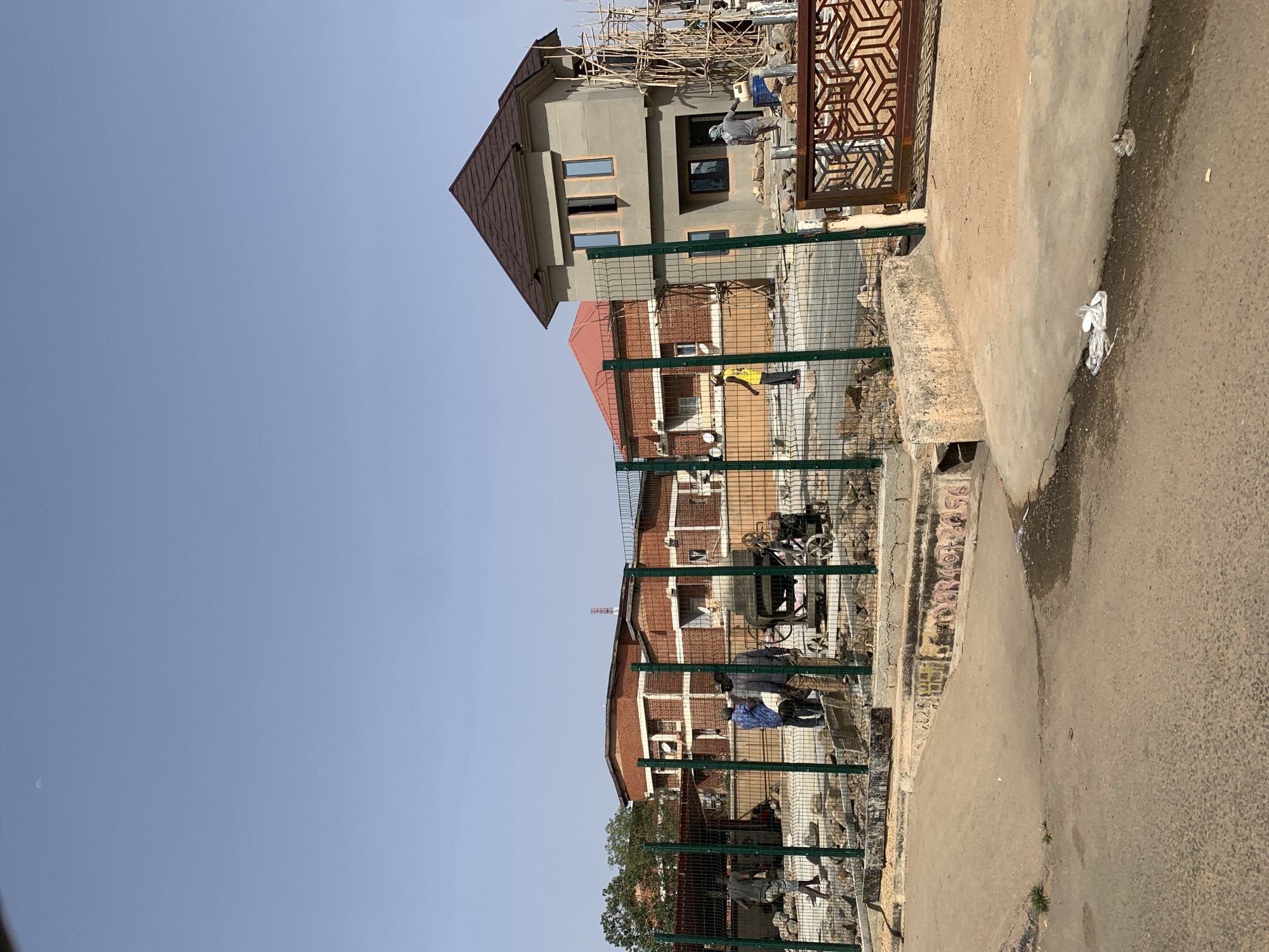 The scene depicts a street in Barakin Lerawi, Nigeria. Visible are several buildings under construction or renovation. One building has a brown roof and gray exterior with scaffolding. Another building is brick with a red roof. A green fence runs along the edge of the property, separating it from the street. People are visible working on the buildings, some wearing blue shirts and another in a yellow shirt. Construction equipment is present behind the fence. The street itself is paved and there is trash or debris on the side. The sky is clear and blue.