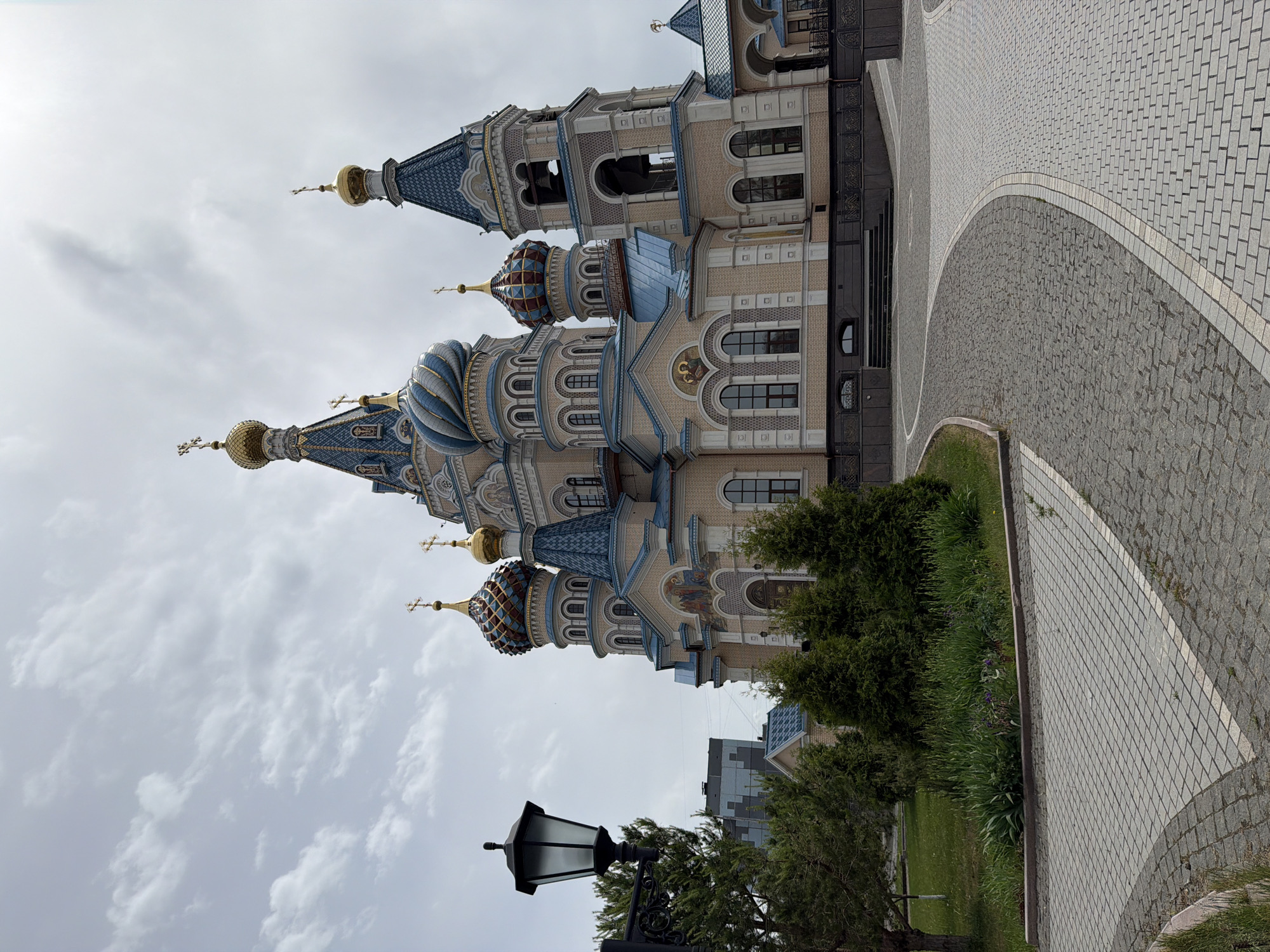 The image shows the Cathedral of Archangel Michael in Izhevsk, Russia. It is a large, ornate building with multiple domes and towers, featuring blue and gold accents. The cathedral is constructed of beige and grey stone with arched windows and decorative details. In front of the cathedral is a paved walkway made of grey and white brick. Adjacent to the walkway is a green lawn with shrubs and a vintage-style street lamp. The sky is overcast with grey clouds. There are no people visible in the image.