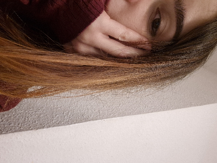 An eye and brow are visible in the top right. A hand is partially covering the face, with fingers near the eye. Long, straight hair with brown and red tones flows across the frame, partially obscuring the hand. A person is wearing a burgundy-colored top. The background transitions from a textured gray surface to a smooth, white one at the bottom of the image.