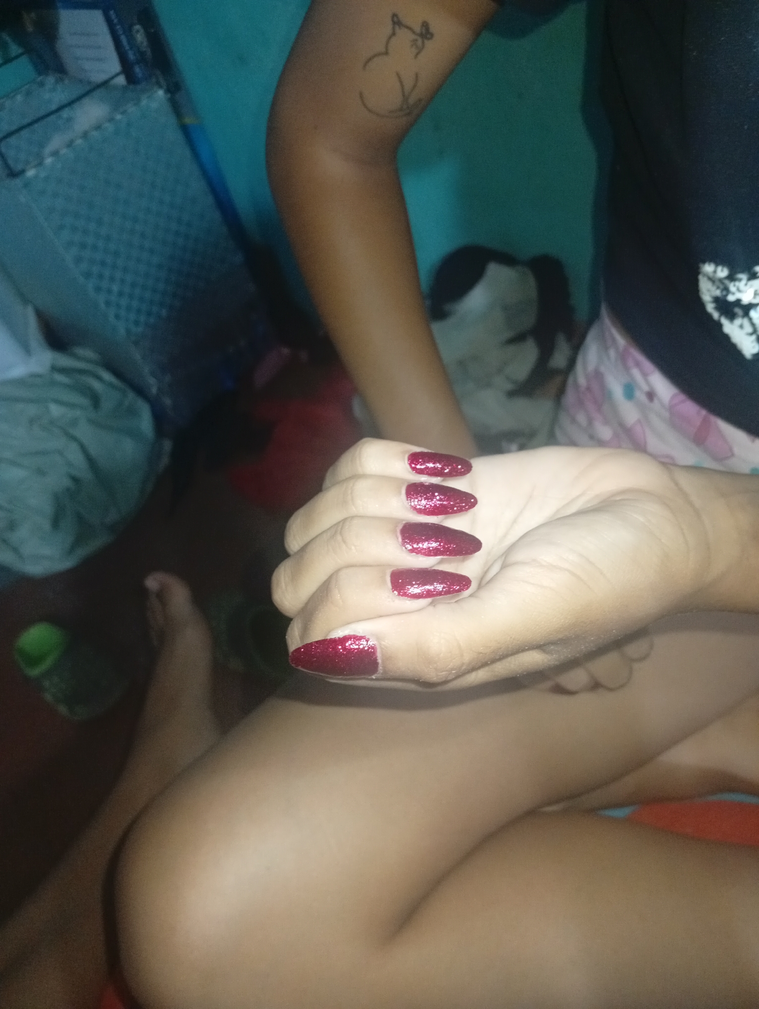 A person sits in what appears to be an indoor environment. Their right arm is prominently displayed, showcasing a tattoo of a cat on their upper arm. The person is wearing a black top and light-colored shorts with a pattern. Their fingernails are painted a dark red color with glitter. Their left leg is visible in the lower-left corner of the image. In the background, there is a blue storage container, a crumpled piece of white fabric, and a framed picture of a person. The specific location is not possible to determine.