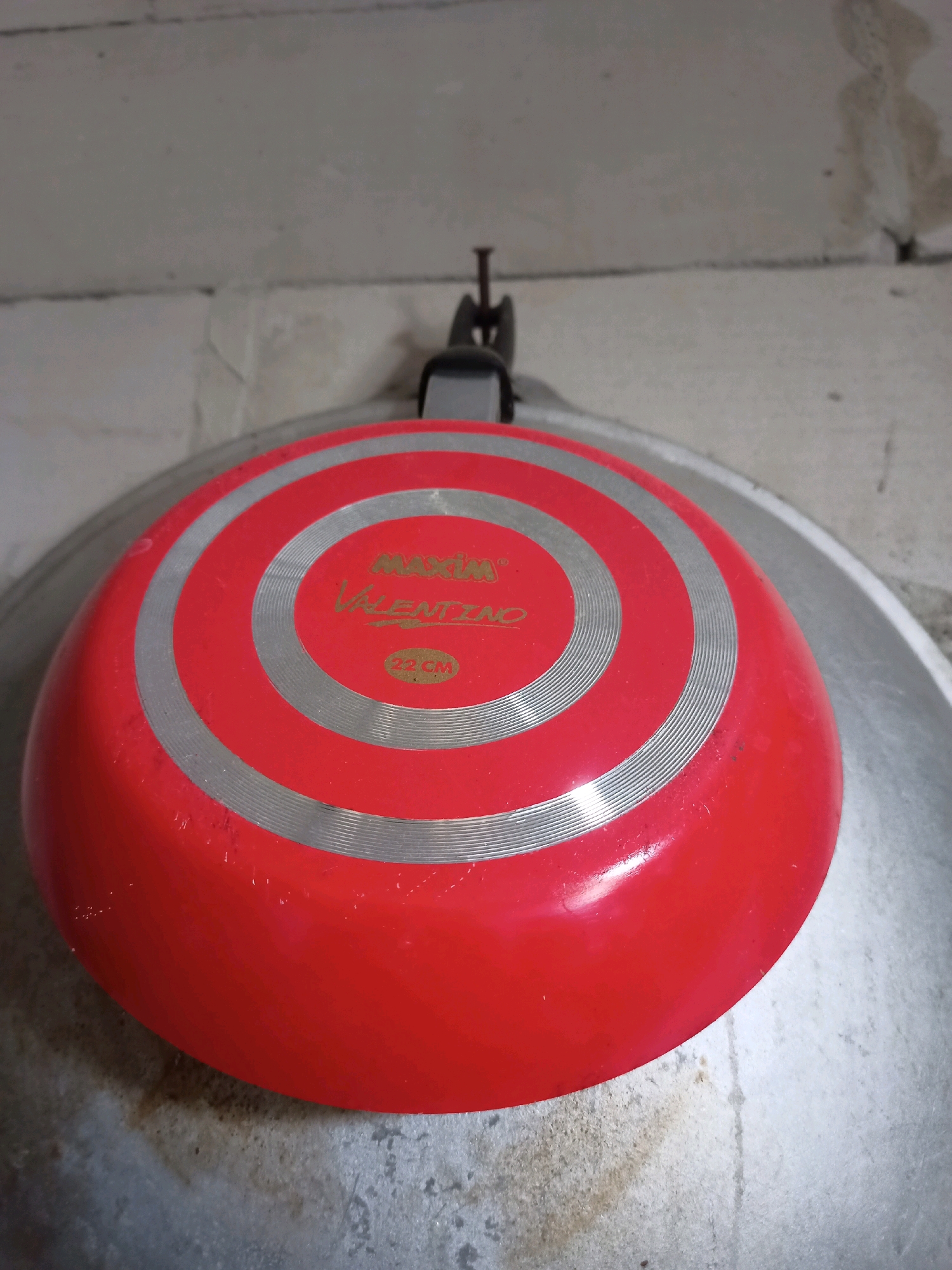A red frying pan with silver concentric circles on its base is hanging from a nail driven into a concrete block wall. A label on the base of the pan reads "MAXIMA VALENTINO 22 CM". The frying pan is resting on top of a larger, metallic pot or pan. The concrete wall appears to be an unfinished interior wall, with visible seams between blocks and some staining. The scene is lit by overhead lighting, casting shadows. The image was taken in the city of Karangtalun, Indonesia.