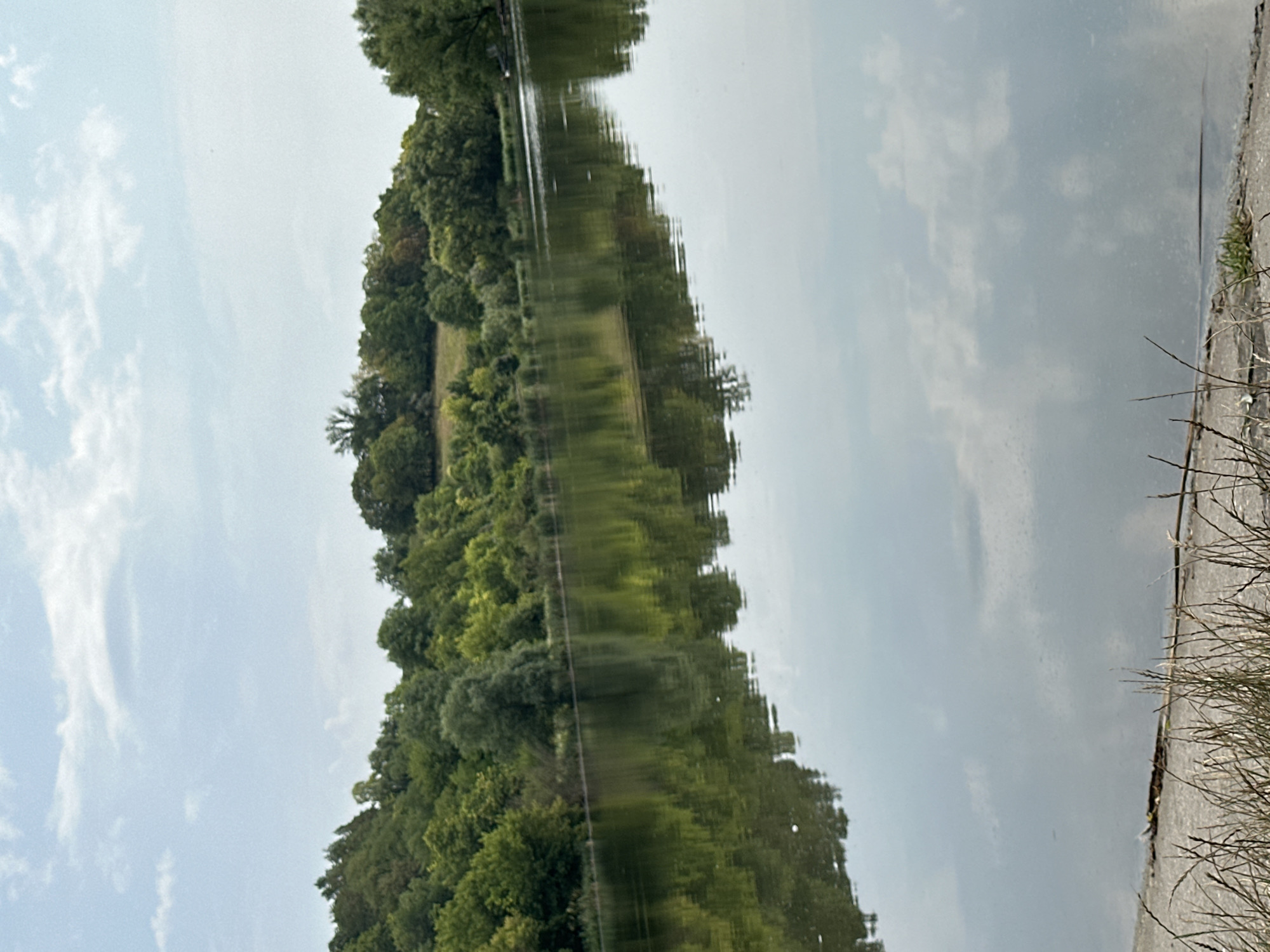 The image depicts a tranquil lake scene in Makiv, Ukraine. The foreground shows a concrete embankment covered with dry grass and reeds. The calm, reflective surface of the lake mirrors the lush green trees and vegetation that line its banks. Beyond the trees, a gently sloping hill is visible, topped with more trees and a clear sky with scattered clouds. The overall atmosphere is serene and natural.