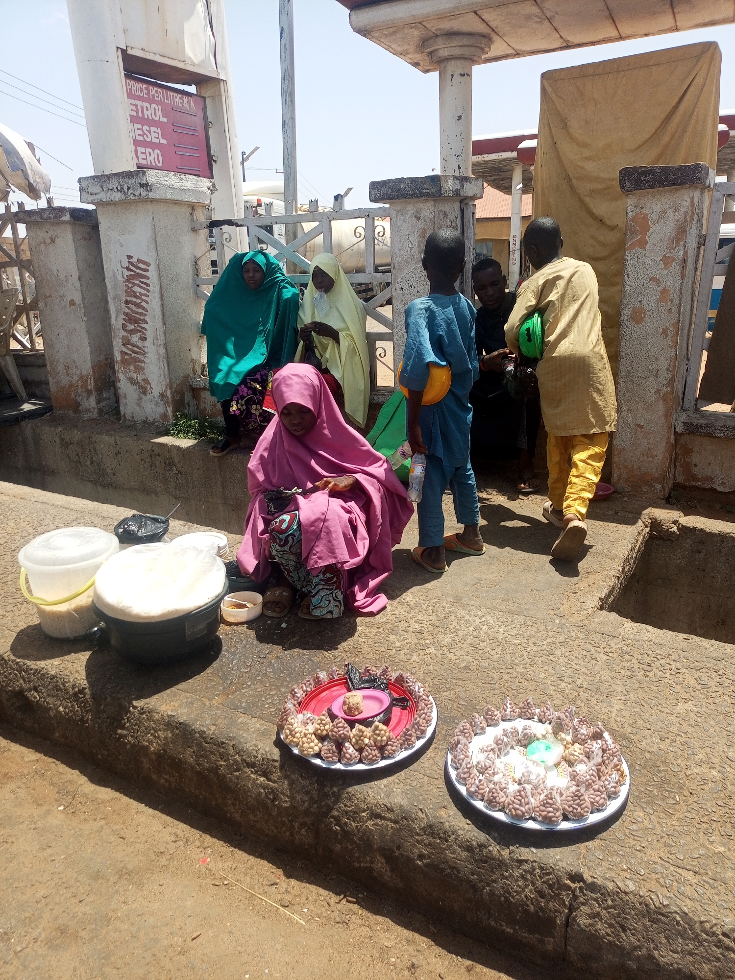 This outdoor image from Gombe, Nigeria, captures a bustling street scene, likely near a petrol station, under bright, clear daylight. In the foreground, a woman wearing a pink hijab and a patterned skirt sits on the rough concrete ground, appearing to be a street vendor. Around her are various food items: two large, round trays filled with individually bagged snacks, possibly groundnuts or candies, a black pot with a white lid, a white bucket containing grains, and a small white bowl.
Behind her, a group of people are gathered. Two women in hijabs, one green and one light yellow, are partially visible. Further back, three young men or boys stand; one in blue holds a plastic bottle, another in light yellow/gold carries a green helmet, and a third in black is less distinct.
The setting is characterized by concrete pillars supporting a canopy structure. One prominent pillar displays a red sign with white text, clearly listing "PRICE PER LITRE," "PETROL," "DIESEL," and "KERO." Vertically painted on an adjacent pillar is the safety warning "NO SMOKING." A metal fence and parts of what appear to be fuel pumps are visible in the background, confirming the petrol station environment. The ground is a mix of concrete and dirt, indicating a well-used public space. The scene conveys a sense of everyday commerce and activity under a sunny sky.
