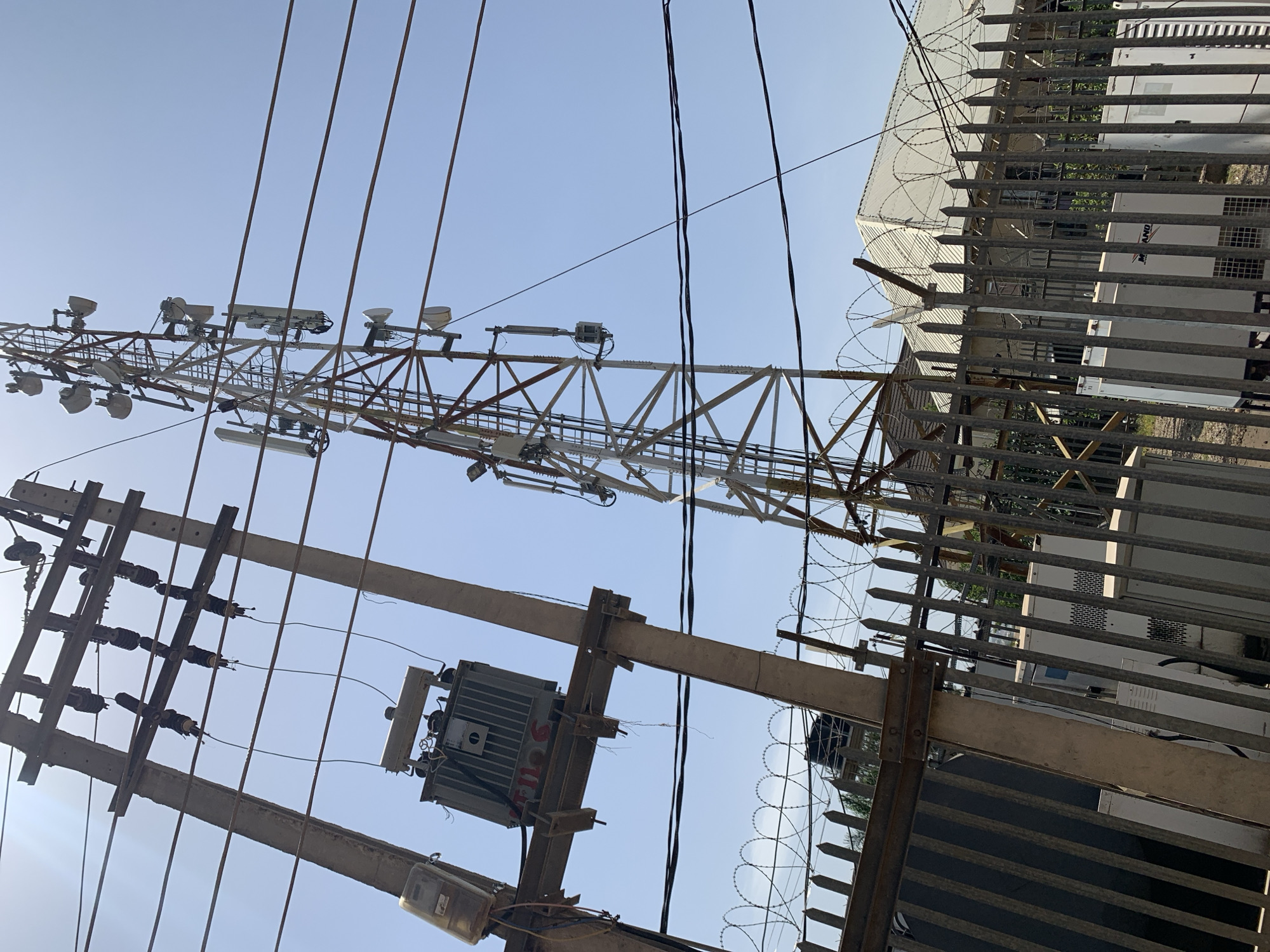 The image shows a scene in Rafin Par, Nigeria. The main elements are utility infrastructure. A tall metal tower stands in the background, equipped with multiple antennas and lighting fixtures. In the foreground is a utility pole with electrical wires and a transformer. To the right of the pole is a metal fence topped with barbed wire, obscuring a white building with mechanical equipment visible through the fence. The sky is clear and light blue.