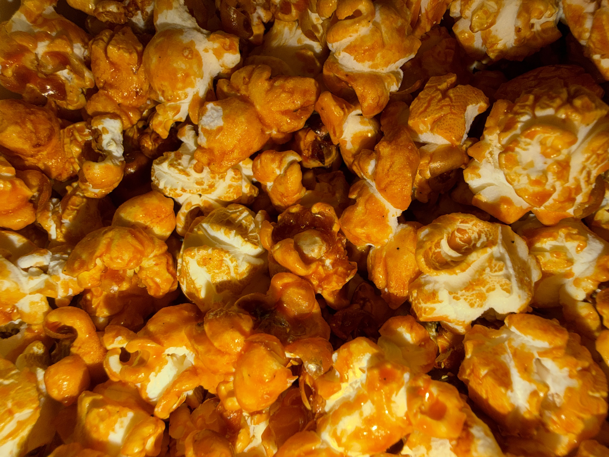 A close-up shot of caramel corn fills the frame. The individual pieces are clusters of popped corn coated in a thick, golden-brown caramel. The caramel glaze varies in thickness and coverage, with some pieces appearing glossy and heavily coated, while others retain more of the popcorn's original white color. The texture appears crunchy and sticky. The shot is tightly cropped, providing a sense of abundance.