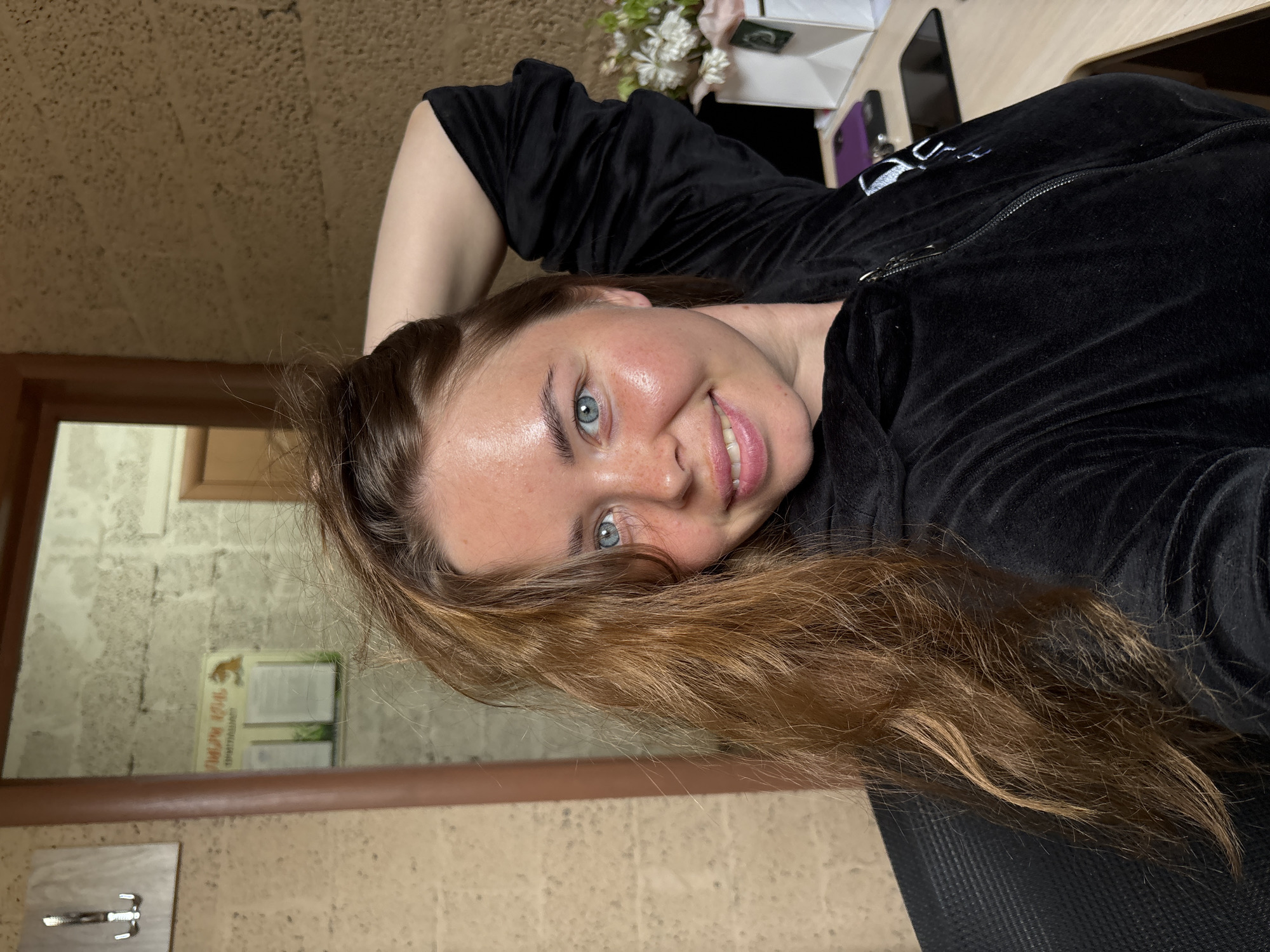 A fair-skinned woman with light blue eyes and reddish-brown hair smiles at the camera. She is wearing a black velvet zip-up jacket. Her right arm is raised above her head. The background shows a room with a tiled wall and a doorway leading to another room. A mirror reflects a wall and a sign in Cyrillic. The scene appears to be indoors. The specific location is Русский Вожой, Russia, as indicated in the instructions. Other objects visible include a table, a phone, and a small floral arrangement.
