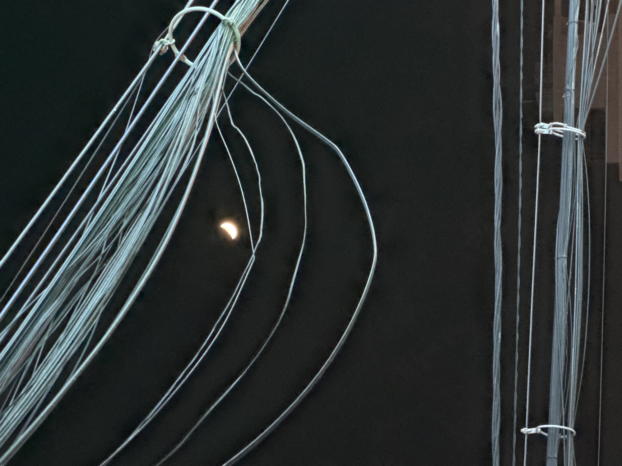 The image shows a crescent moon visible through a dense network of overhead electrical wires against a dark sky. The wires are arranged in multiple layers, some tightly bundled and others draped loosely, forming a chaotic urban landscape. The moon, illuminated by sunlight from behind, appears as a bright sliver against the blackness. The lower portion of the image reveals additional layers of wires and what appears to be the edge of a building or structure. The scene is likely an urban setting in Nha Trang, Vietnam, characterized by its ubiquitous overhead wiring. There are no people or discernible activity depicted, focusing solely on the celestial event framed by the man-made infrastructure.