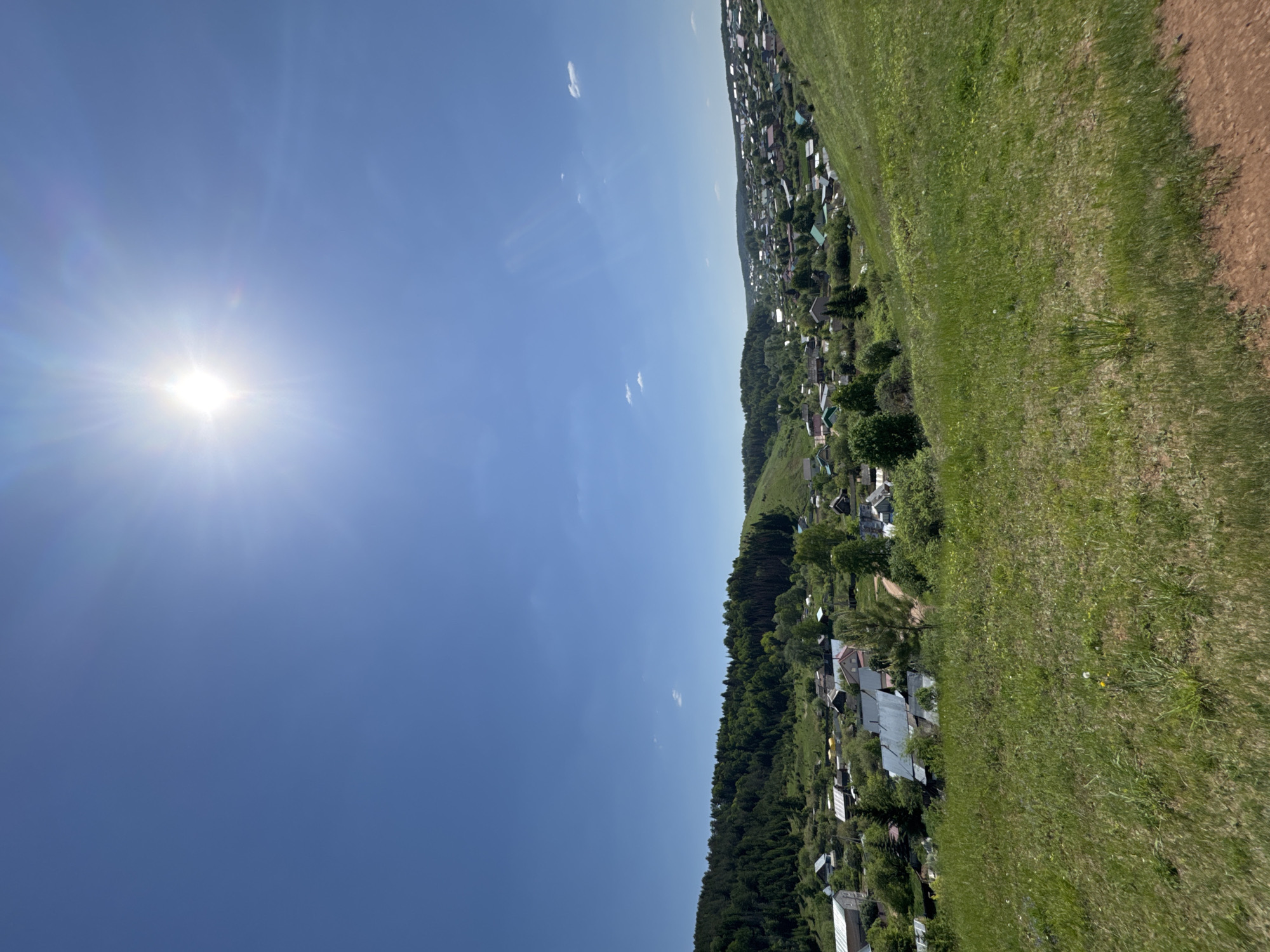 The image is an aerial shot of Ольховка, Russia, taken on a sunny day. The sun is prominent in the upper left, shining brightly in a clear blue sky with a few scattered clouds. A strip of greenery, likely a field, runs along the right side of the frame. A dirt path borders the grassy area. To the left of the field is a landscape of trees and residential buildings, giving the appearance of a dense village. The houses feature a variety of roofing styles.