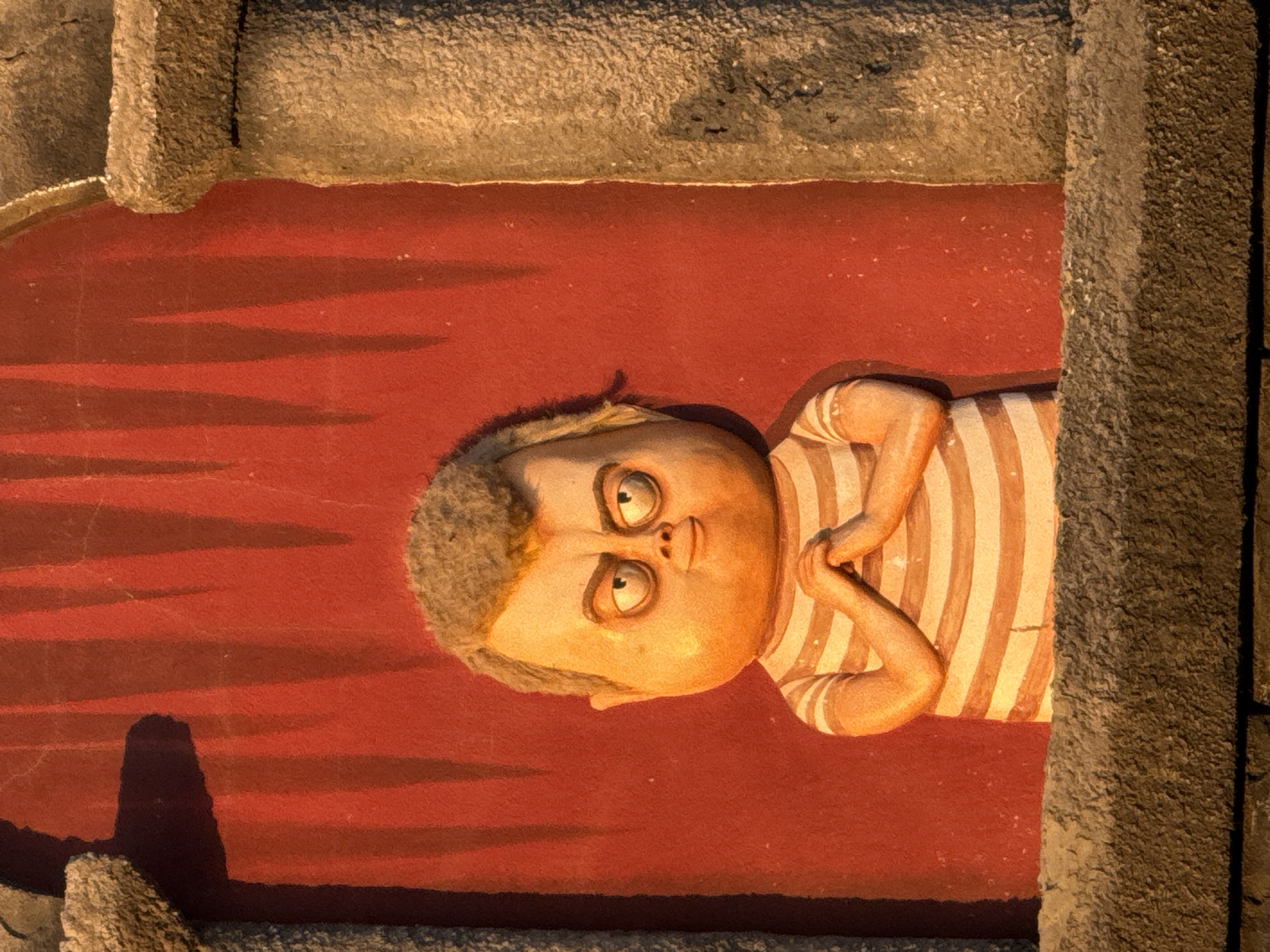 This image shows a close-up, low-angle, medium shot of a statue of a character with a round, pale face, grey, curly hair, and wide, staring eyes. The character is wearing a striped shirt and has its hands clasped together in front of its chest. The statue is positioned in front of a textured, bright red background with dark, vertical shadow lines. To the left and right of the statue are grey, rough-textured architectural elements, possibly framing the scene. The lighting appears to be warm and directional, casting shadows on the background and highlighting the texture of the statue and architectural elements. The image does not provide any information about its location in Novofedorovka, Ukraine.