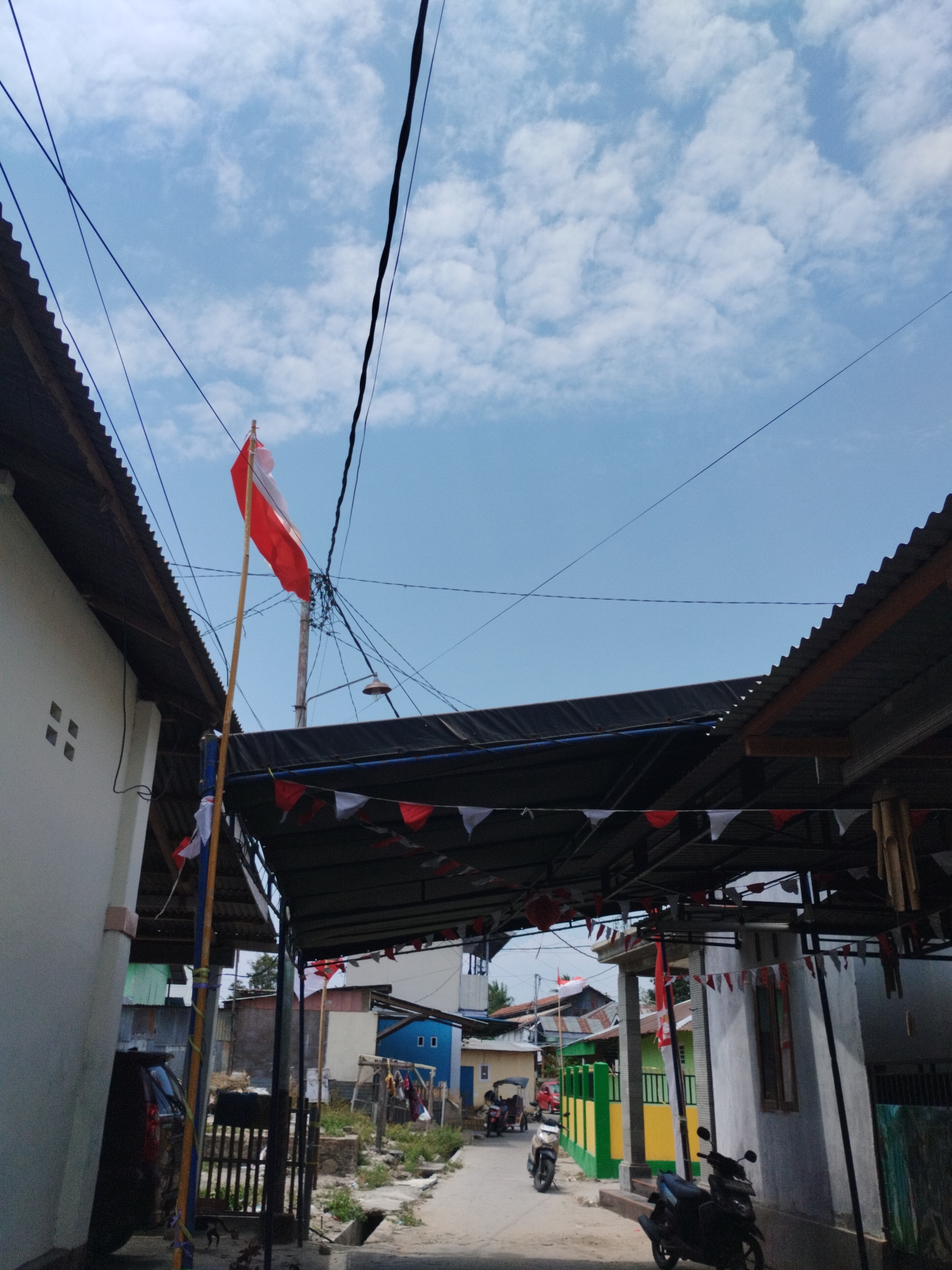 The image depicts a street scene in a residential area, likely in Gorontalo, Indonesia, given the presence of the Indonesian flag (red and white horizontal bands) and celebratory bunting. The Indonesian flag is prominently displayed on a tall wooden pole on the left side of the image.
The street appears to be paved, with a drainage ditch running along its right side. Buildings line both sides of the street. On the left, a white building with a corrugated metal roof is visible. A dark-colored SUV is parked in front of a wooden fence. On the right side, a series of buildings with corrugated metal roofs and colorful painted walls (green and yellow are noticeable) are present. A covered walkway or awning, decorated with red and white triangular flags, extends from one of the buildings.
Several motorcycles are parked along the street, and two appear to be in motion in the background. The sky is blue with scattered white clouds, suggesting a daytime scene. Power lines are strung across the sky, contributing to the urban or semi-urban environment. The overall atmosphere suggests a local community, possibly preparing for or celebrating a national holiday.