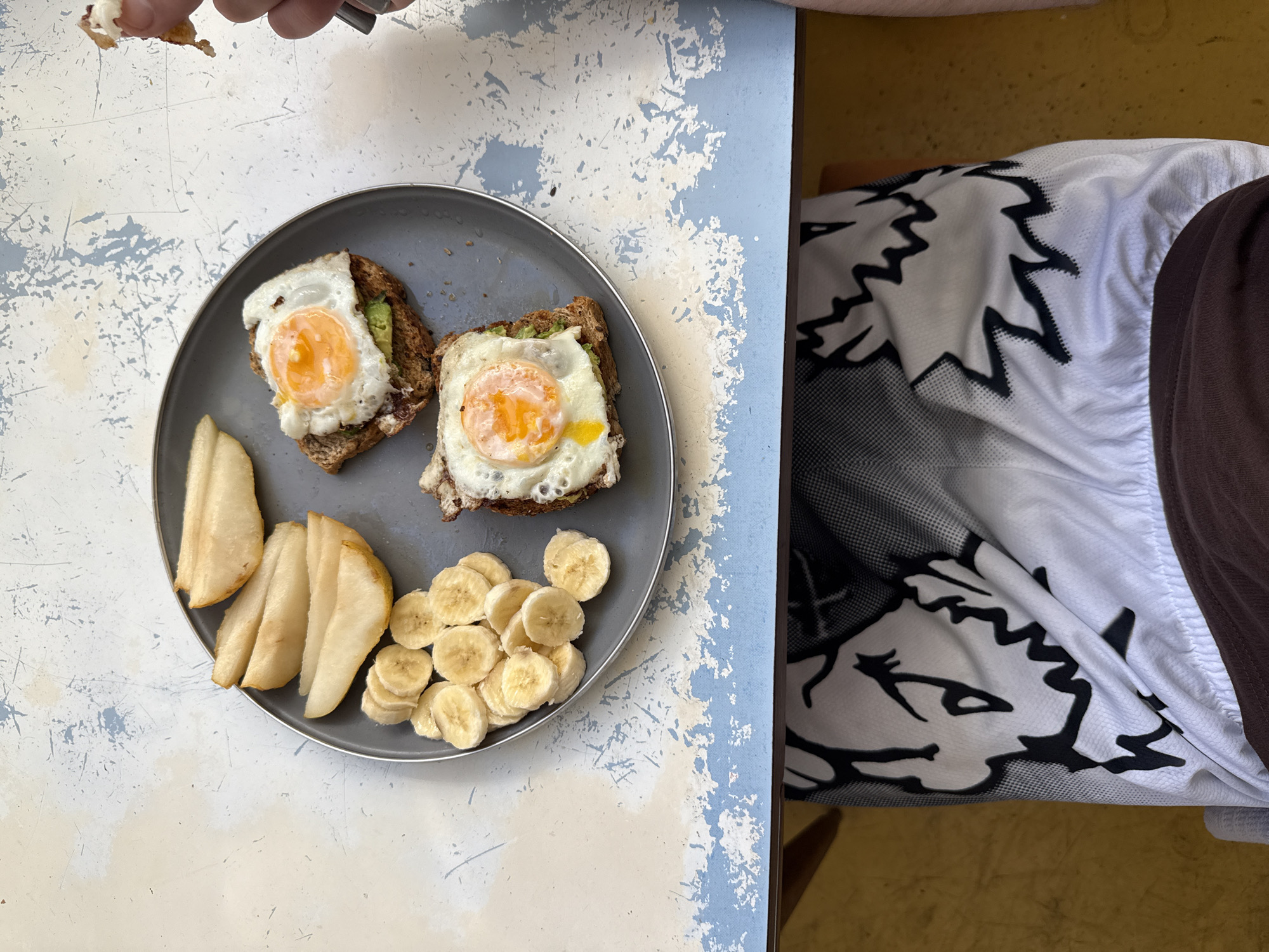 An overhead shot captures a casual breakfast or brunch setting on a distressed light blue and white table. A grey, round plate prominently features two pieces of toast, each topped with a perfectly fried egg and mashed avocado. Accompanying the toast are several slices of pear and a neat pile of sliced bananas.
In the upper left corner, a person's hands are visible, holding a small piece of food or perhaps manipulating it with a partially seen metal utensil. To the right side of the frame, another person's body or leg is visible, clad in a white garment with a bold black, jagged pattern that resembles a stylized fox head, suggesting activewear or a sports jersey. This is paired with brown shorts or pants. The background beyond the table appears to be a warm, yellowish-brown surface, likely an interior wall or floor. The scene depicts a relaxed mealtime, characteristic of an everyday moment in a home or cafe, set in Buenos Aires, Argentina. No discernible text is visible in the image.