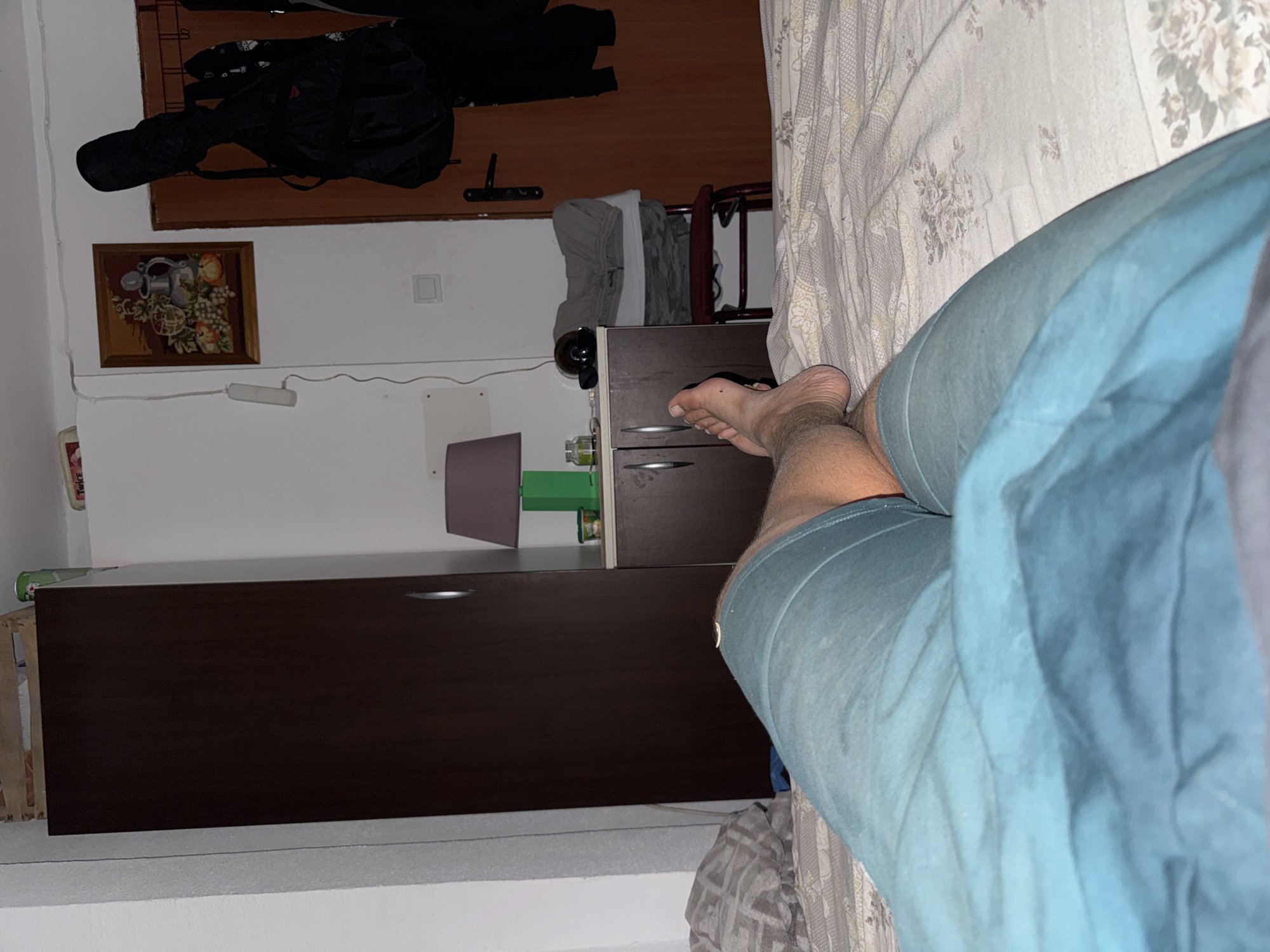 The image depicts a dimly lit bedroom. In the foreground, a person's legs, clad in blue shorts, are visible, resting on a bed with a floral patterned duvet. A bare foot is visible to the right. In the background, a dark brown wardrobe stands against a white wall. To the right of the wardrobe is a small cabinet with a purple lamp and some containers on top. A framed tapestry depicting fruit hangs on the wall above the cabinet. To the far right, a wooden door with a handle is visible, with a black guitar case and several coats hanging on the wall beside it. A small red chair with folded clothes is positioned in front of the door. The overall impression is that of a lived-in, personal space. The location is specified as a bedroom in Sutomore, Montenegro.