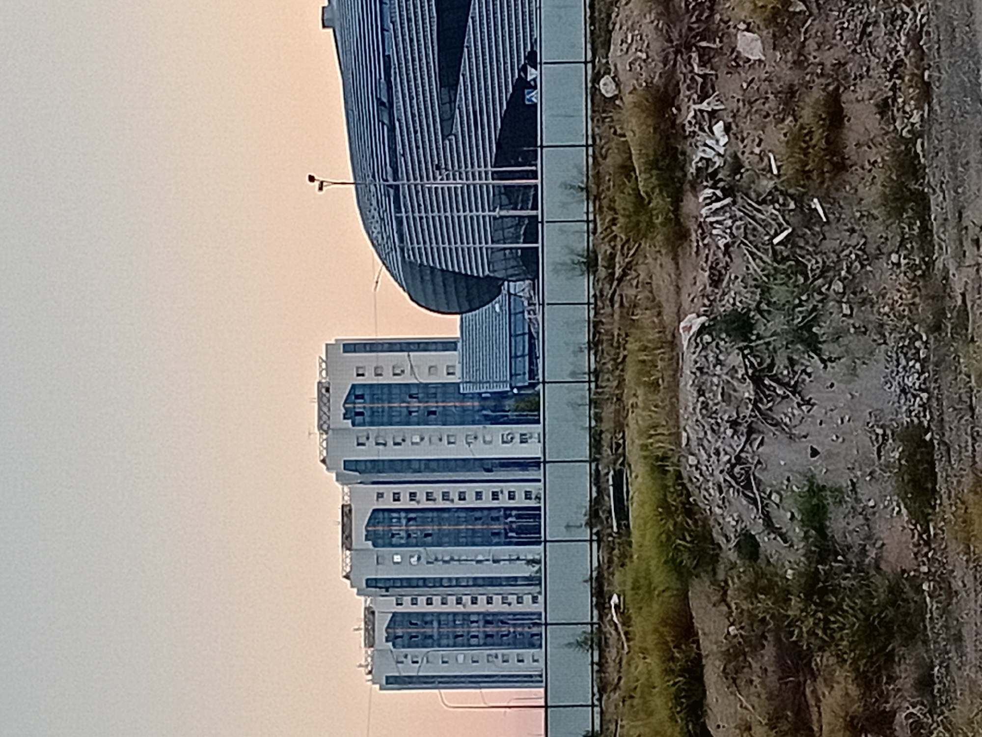 The image shows a contemporary urban landscape in Semey, Kazakhstan, featuring several modern residential buildings and a distinctive, curved architectural structure. The residential buildings are tall and appear to be composed of light-colored material with dark blue, glass-fronted sections, arranged in a row. To the right of the residential buildings is the curved structure, characterized by its metallic, ribbed facade and large glass panels at its base. A street lamp with two fixtures stands in front of this structure. The foreground of the image reveals a rugged, unkempt terrain with sparse vegetation, gravel, and debris, suggesting a construction site or undeveloped land. A low fence separates the foreground from the background buildings. The sky above is a pale gradient of orange and blue, indicative of either sunrise or sunset. No people are visible in the frame.