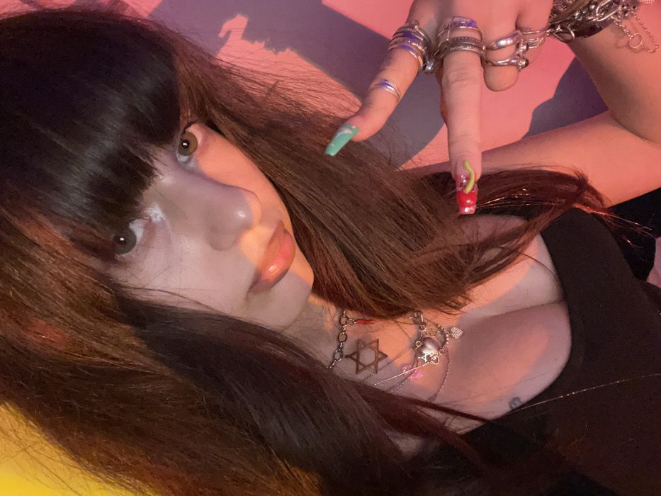 A person with dark brown hair and bangs poses at an angle, showcasing several rings on their fingers and colorful nails. They wear a black top, and several necklaces are visible, one featuring a Star of David. The lighting is a mix of pink and yellow hues, adding a vibrant touch to the composition. The person's face is prominent, with a slight upward gaze.