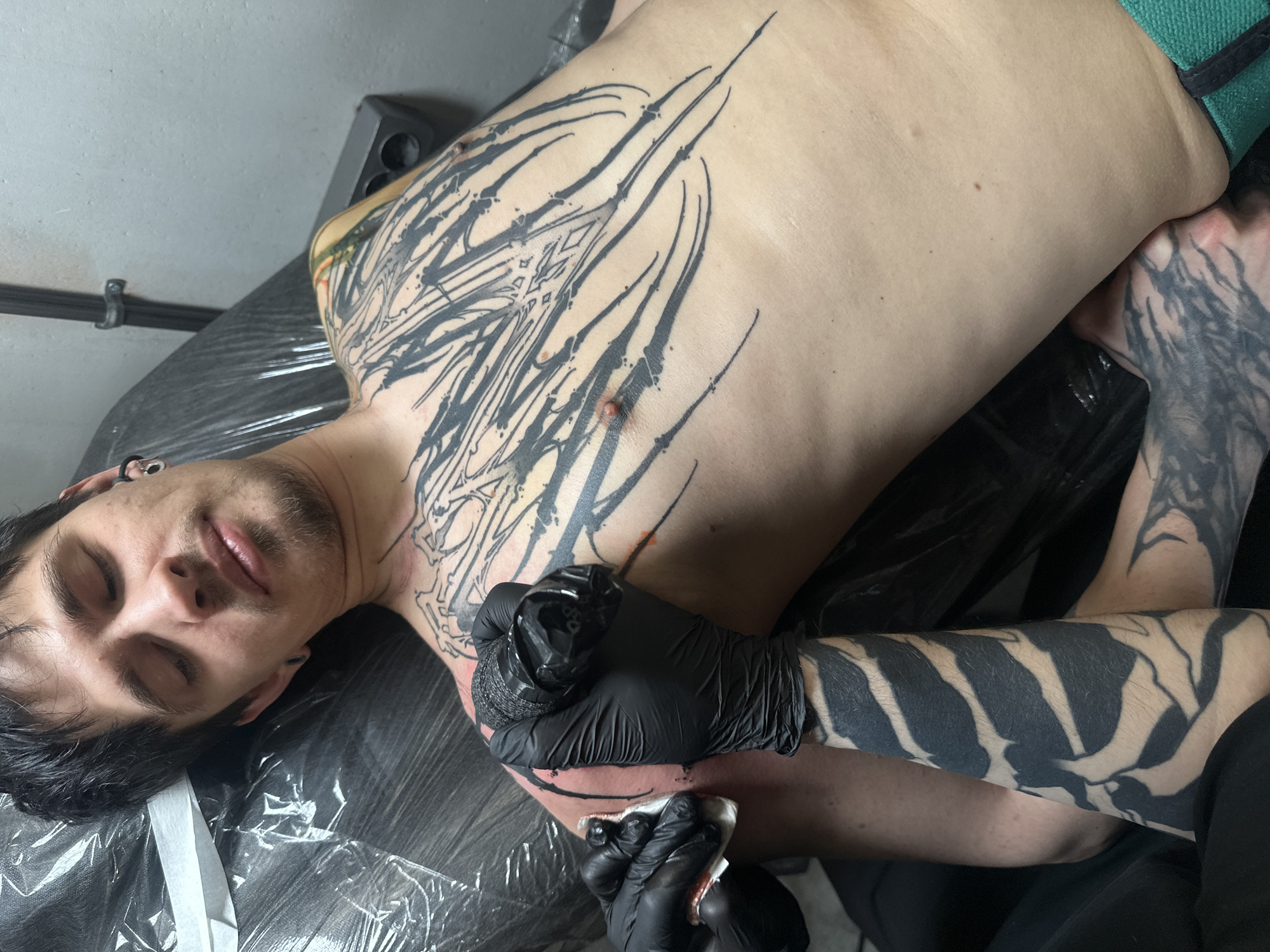 Here is a detailed description of the image:
**Setting:**
The image appears to be taken inside a tattoo studio. The visible background is simple, with a gray wall and some equipment.
**People:**
* One person is lying down, receiving a tattoo on their upper torso, which is visible. They have dark hair, fair skin, and visible ear piercings.
* Another person, presumably the tattoo artist, is working on the tattoo, with their hands visible. They are wearing black gloves.
**Subjects:**
* A new tattoo is being applied to the person's chest and upper torso. The design is primarily black and appears complex, with lines and abstract shapes.
* The person also has existing tattoos on their arm/shoulder.
**Objects:**
* Tattooing equipment, including the tattoo machine, is being held by the artist.
* A disposable wipe is held to clean excess ink.
* The person is lying on a table covered with a black plastic sheet.
**Actions:**
* The tattoo artist is in the process of applying the tattoo.
* The person is lying still, receiving the tattoo.
**Interactions:**
* The tattoo artist is directly interacting with the person's skin.
**Location:**
The location cannot be determined using this image.