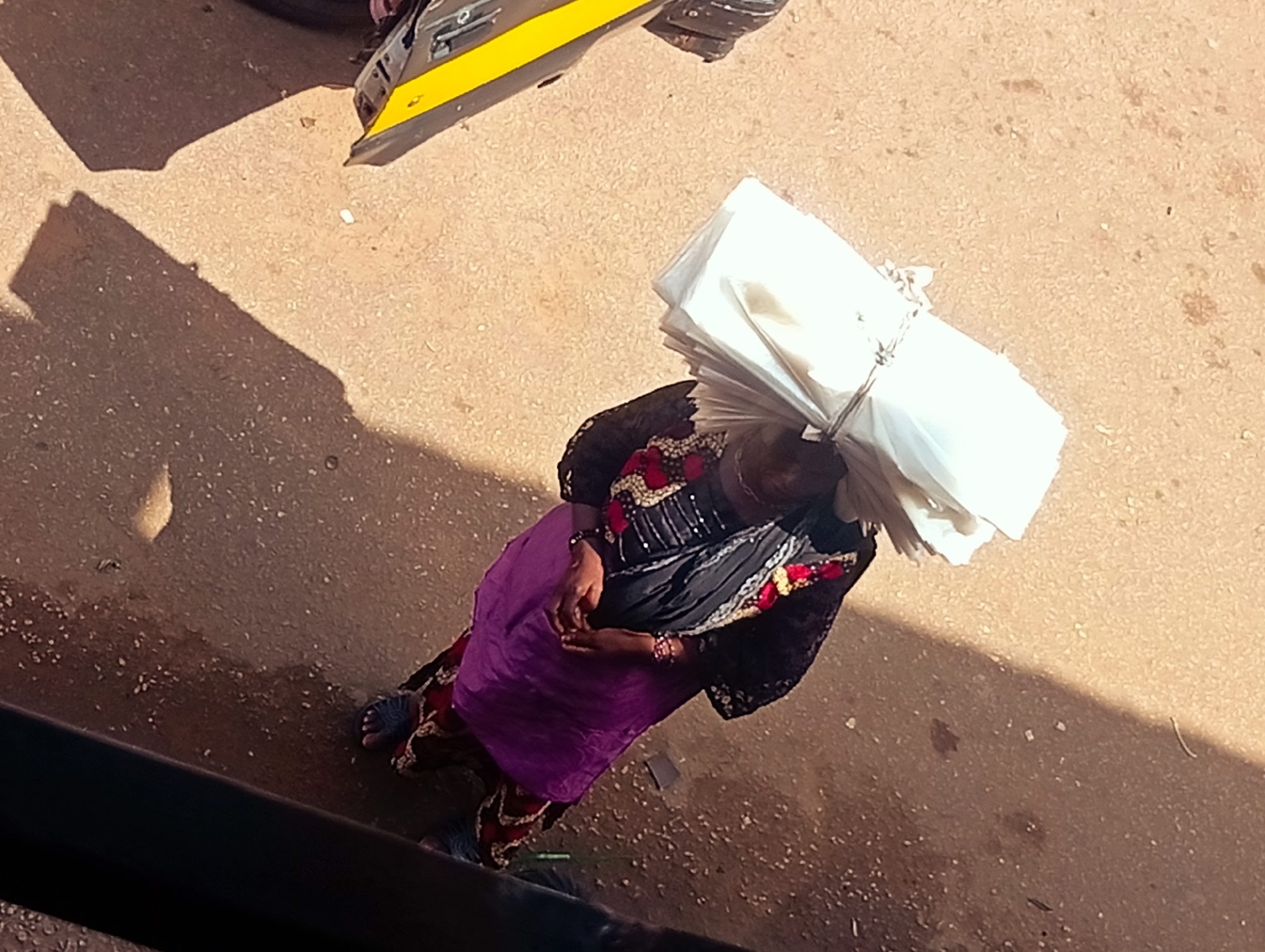 In the city of Bukuru, Nigeria, a person is seen from a high angle, carrying a large stack of white material on their head. The individual is wearing a black garment with ornate red and white embroidery around the neckline and cuffs, and a vibrant purple skirt. Their hands are clasped together in front of them. The ground is a textured, sandy surface, with shadows cast by the person and another object, possibly a vehicle, partially visible at the top of the frame. The lighting suggests it is daytime.