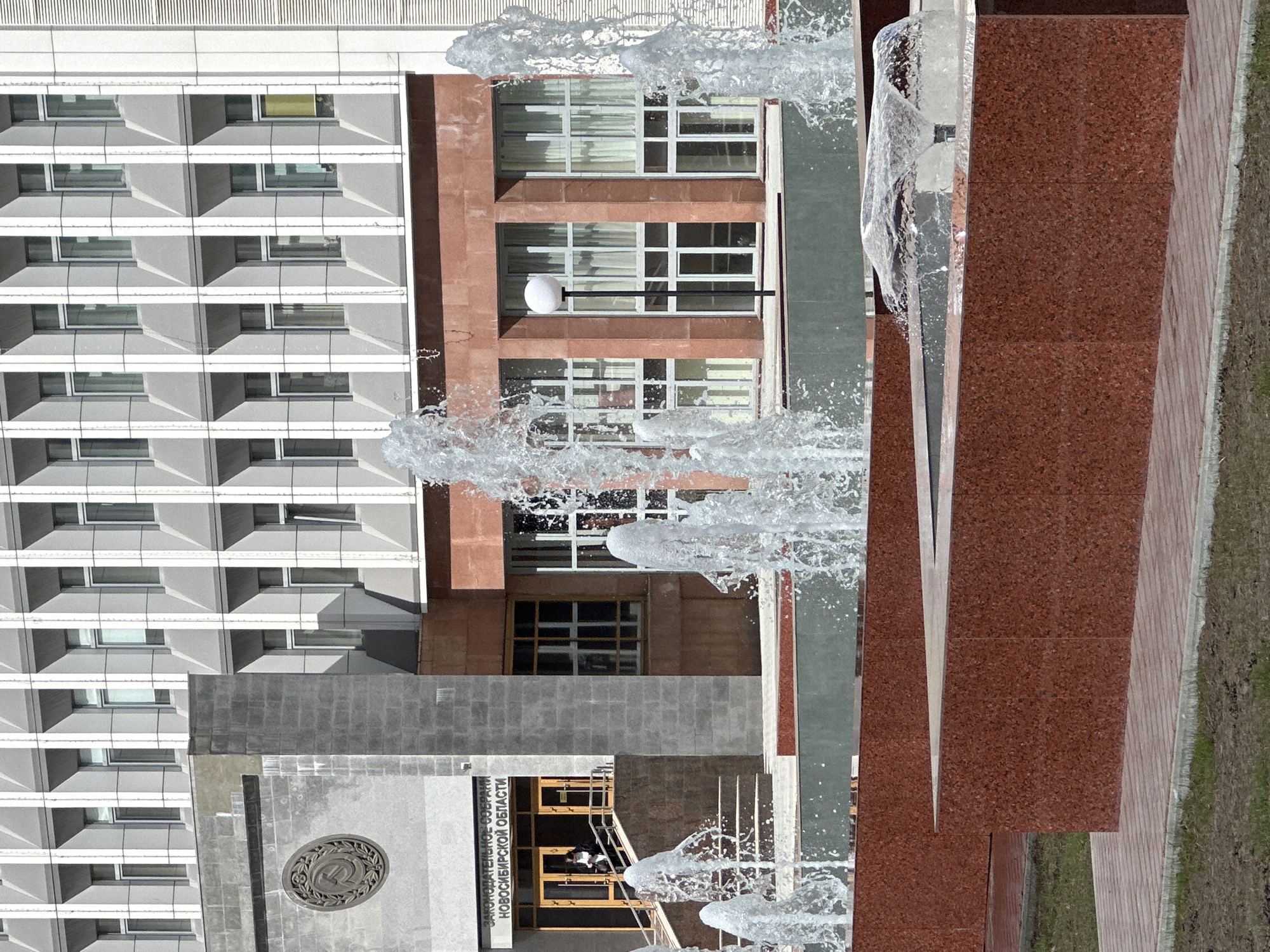 The image shows a partially obscured view of a building complex in Novosibirsk, Russia, likely taken from a low angle due to the presence of a fountain. The buildings consist of a white building with a grid-like facade of windows, an orange brick building with a similar grid-like window structure, and a lower gray section. The gray section has an emblem and inscription in Russian, which identifies the building as the "LEGISLATIVE ASSEMBLY OF THE NOVOSIBIRSK REGION". In the foreground is a fountain. People are visible near the entrance of the building.
