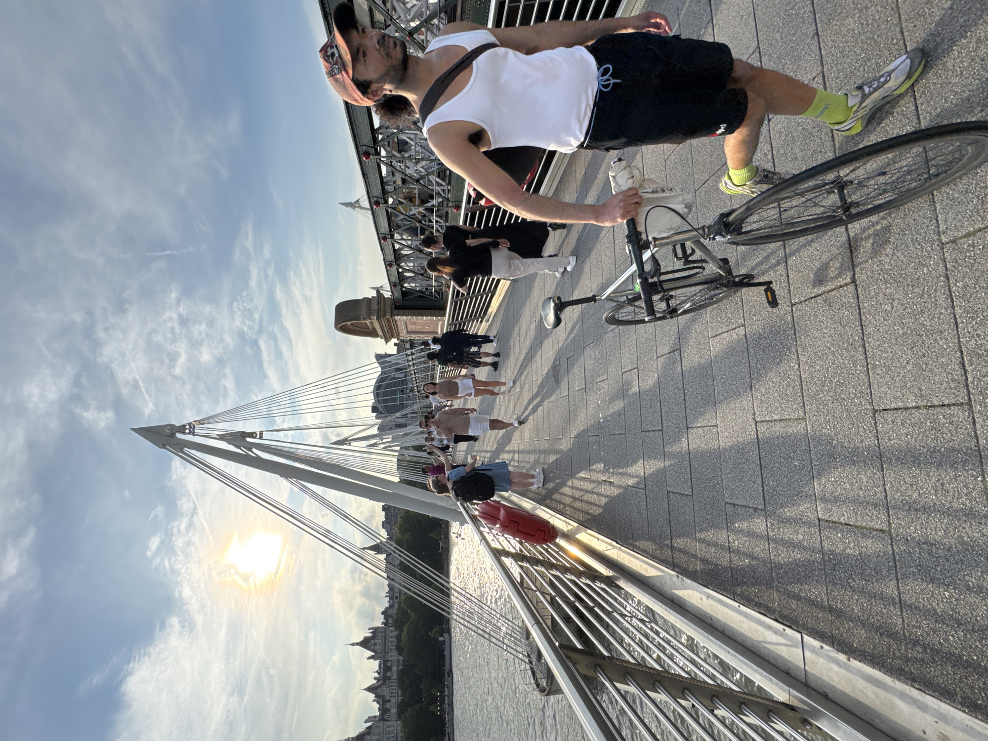 A man wearing a white tank top, black shorts, a patterned baseball cap, and bright yellow socks walks across a pedestrian bridge in London, United Kingdom, holding the handlebars of a silver bicycle. He is walking towards the right of the frame, with the bicycle to his left. The bridge, identified as the Golden Jubilee Bridges, has a modern design with white suspension cables. Several other people are walking in both directions on the bridge. The sun is low in the sky, casting long shadows across the stone-paved walkway. In the background, the River Thames is visible, with buildings and architectural landmarks of London, such as the Houses of Parliament and the London Eye, in the distance. The sky is blue with scattered white clouds.
