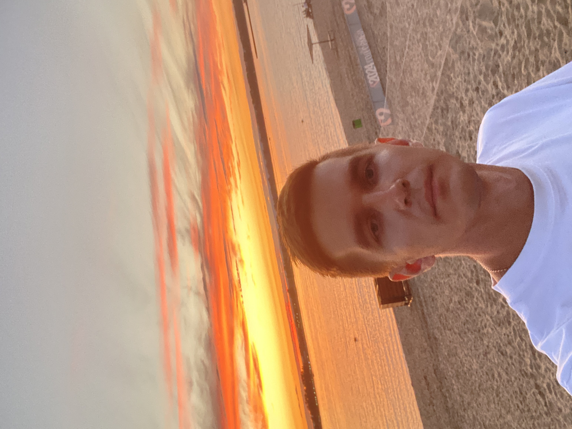 A man with short, light brown hair takes a selfie on a sandy beach during sunset. The sky is filled with vibrant orange and yellow clouds. A body of water stretches across the middle of the image, reflecting the sunset colors. In the background, there is a beach with a few scattered objects, including a small structure, a trash can, and a banner that reads "2024." The man is wearing a white t-shirt and a silver chain. The overall scene suggests a recreational area within or near Novokuybyshevsk, Russia, given the presence of the banner and the commonality of such landscapes.