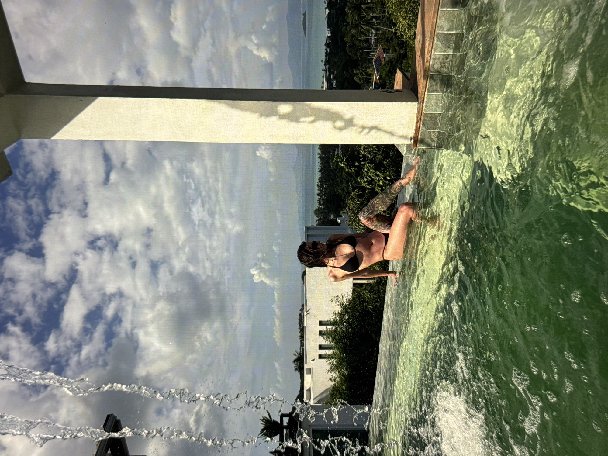 A person in a black bikini sits on the edge of an infinity pool, their legs dangling in the water. They have tattoos on their legs and arms. The pool water is a vibrant green. In the background, there is a lush green landscape with palm trees and white buildings, overlooking a body of water and distant hills. The sky is cloudy with patches of blue. A structure, possibly a pillar, casts a shadow on the pool's edge. Water from a fountain splashes in the foreground. The image appears to be taken during the day, with bright sunlight illuminating the scene.