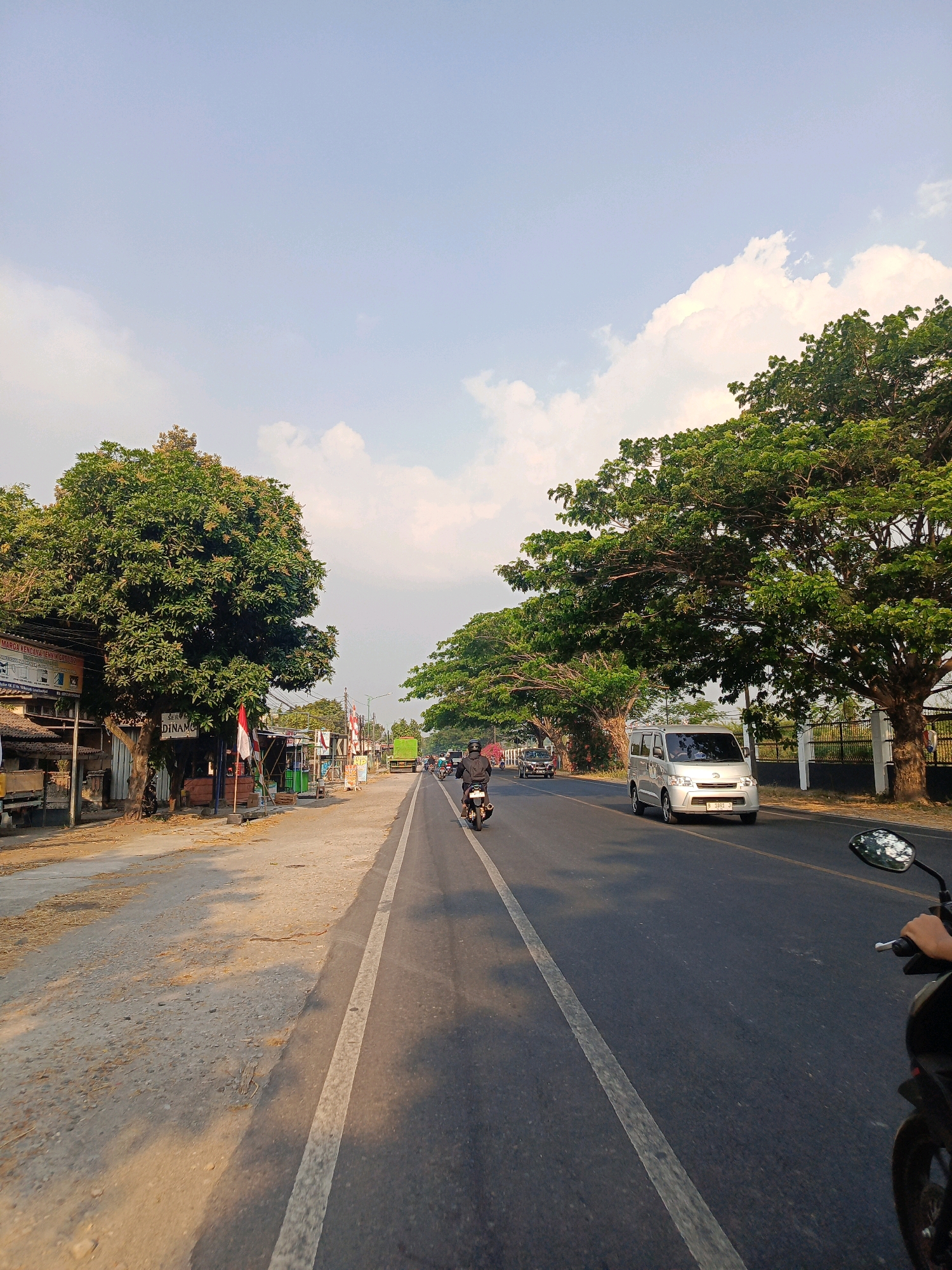 The image depicts a street scene in Maospati, Indonesia, on a bright, clear day with a few clouds. A two-lane asphalt road stretches into the distance, bordered by trees and buildings on the left and trees on the right. Several vehicles are present, including motorcycles, a silver van, and a green truck. A person is riding a motorcycle in the foreground, and another person is riding a motorcycle further down the road. The buildings on the left appear to be shops or small businesses, with some signage visible. The overall atmosphere is that of a typical day in a town or city in Indonesia.