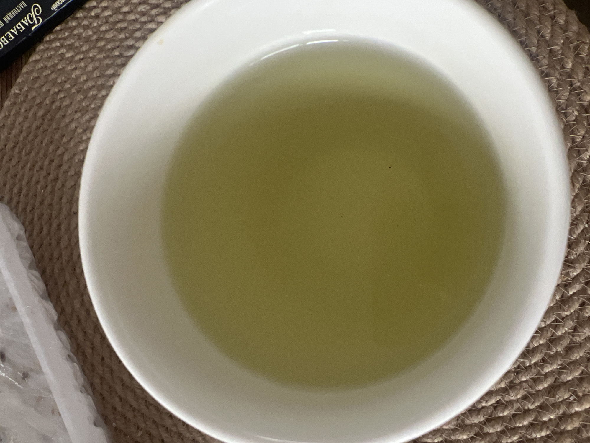 This image is a close-up overhead shot of a white bowl filled with a pale green liquid, likely tea or a herbal infusion. The bowl sits on a brown woven placemat. In the upper left corner, a portion of a white plastic container is visible, containing what appears to be food items. To the right of the bowl, a dark blue box with gold lettering, possibly packaging for tea or medicine, is partially visible. The scene is indoors, and the lighting suggests it is daytime. There are no people or animals present in the image. The image does not contain information to suggest it is located in Votkinsk, Russia.