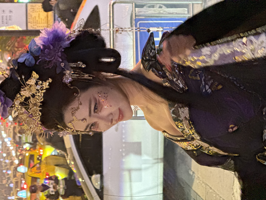 A woman is dressed in an ornate, traditional-style costume with intricate gold headwear adorned with purple flowers and butterflies. Sparkling makeup highlights her eyes and cheeks. She is leaning slightly forward, smiling gently. Behind her, there are blurry lights and vehicles suggesting an urban environment. She is standing next to a white vehicle or structure. The angle is somewhat tilted, giving a dynamic perspective.