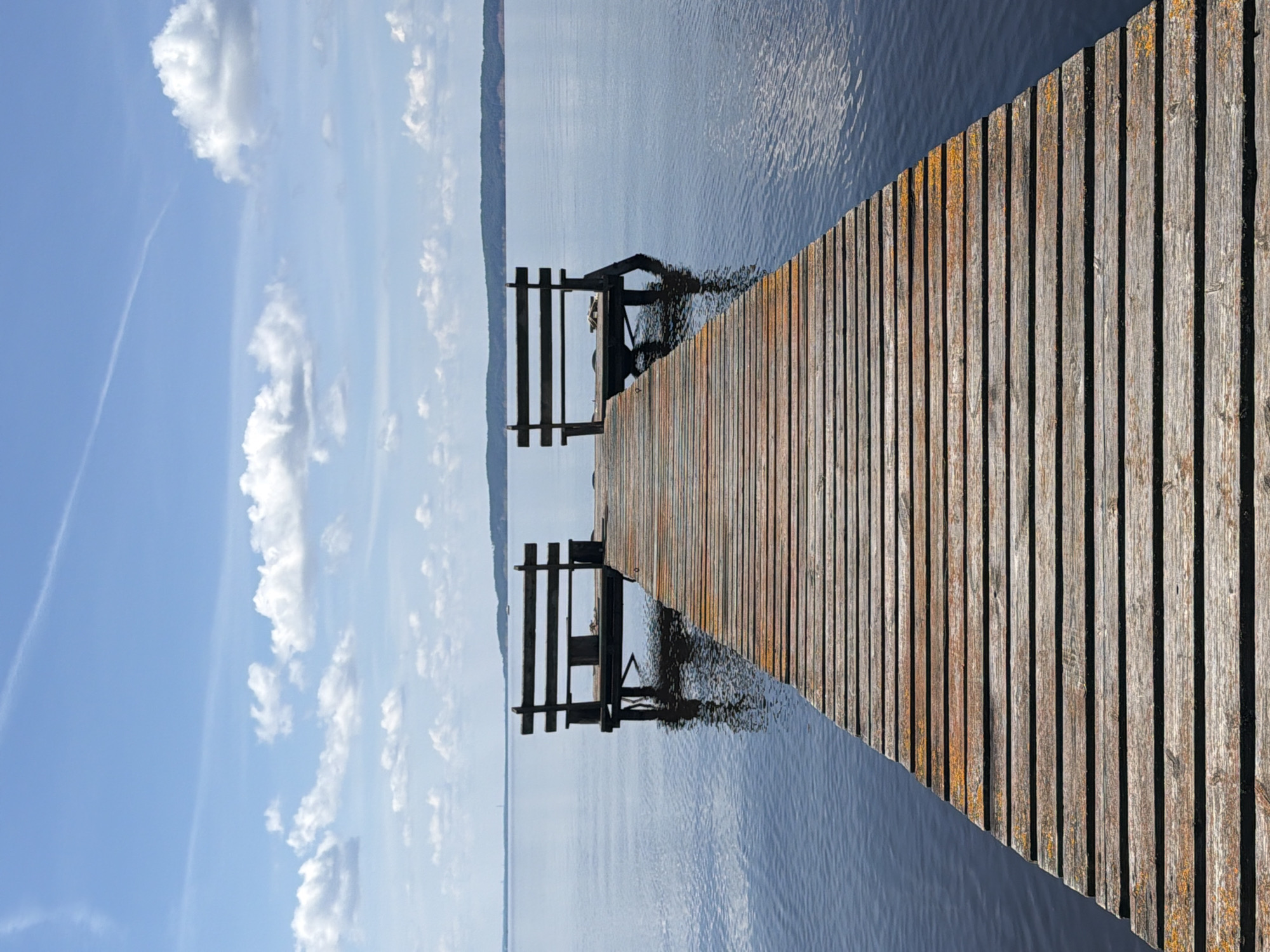 A wooden pier extends into a calm body of water under a bright blue sky with scattered white clouds. Two weathered wooden benches are positioned at the end of the pier, facing the water. The water is dark blue and reflects the sky. A distant, tree-covered shoreline can be seen on the horizon. The scene is peaceful and serene, with no people present. The location is identified as Stepanovo, Russia.