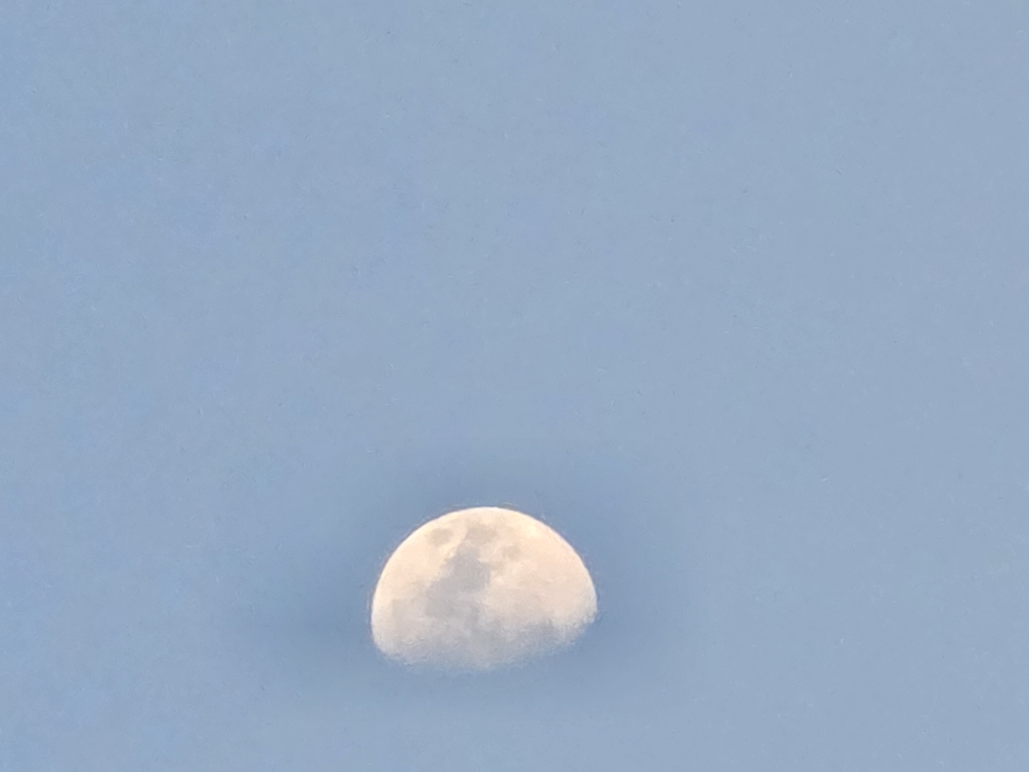 The image shows a partial view of the moon against a light blue sky. The moon appears to be in its first quarter phase, with approximately half of its surface illuminated. The illuminated portion of the moon displays subtle variations in tone, suggesting the presence of craters and lunar maria. The sky is a uniform light blue, with no clouds or other celestial bodies visible. There are no structures, landmarks, or any indication of the city of Yambol, Bulgaria, present in the image.