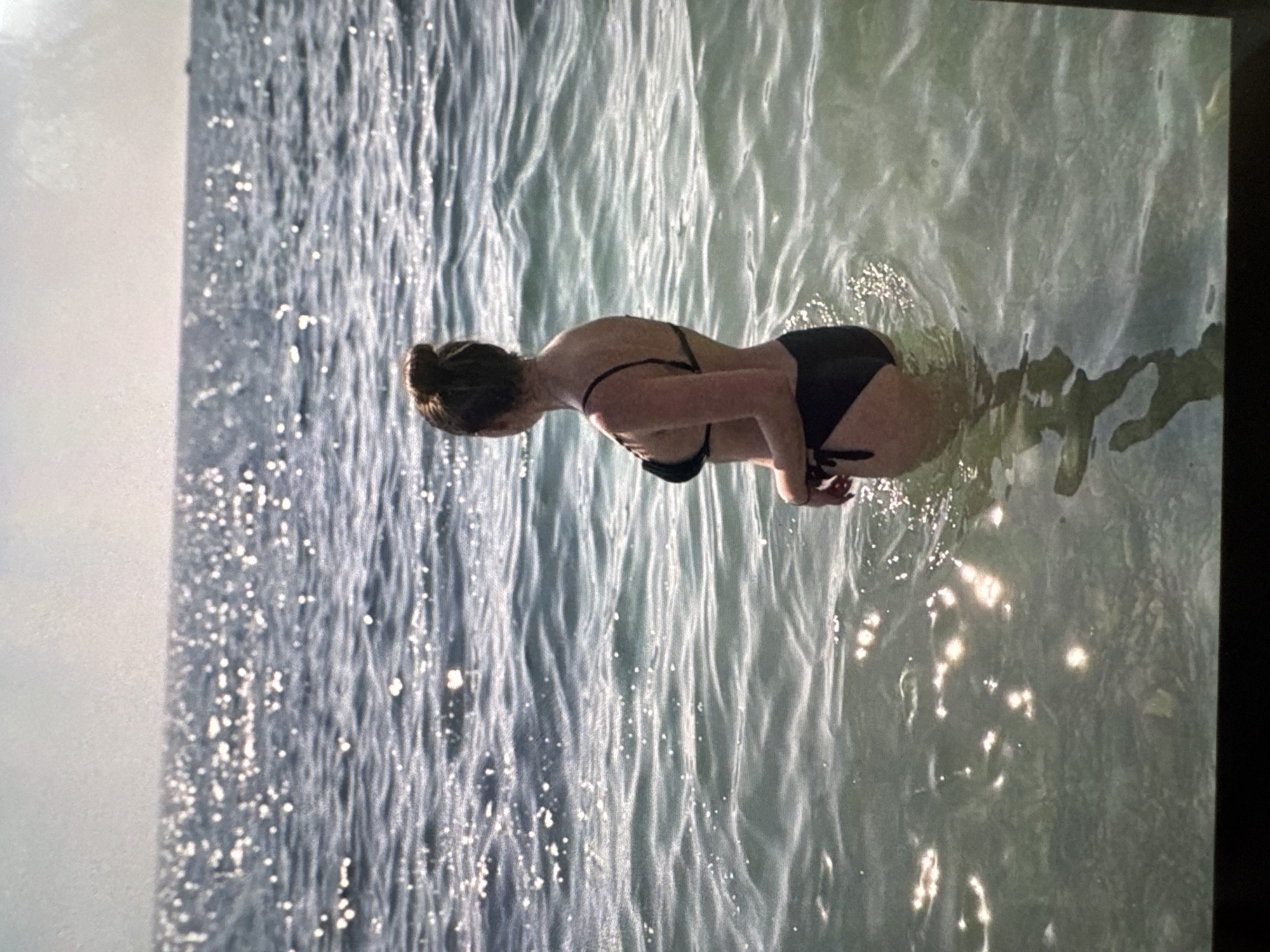 A woman stands in shallow water, facing away from the camera and looking out at the ocean. She is wearing a black bikini. The sun glints off the water, creating a shimmering effect. The horizon is visible in the distance.