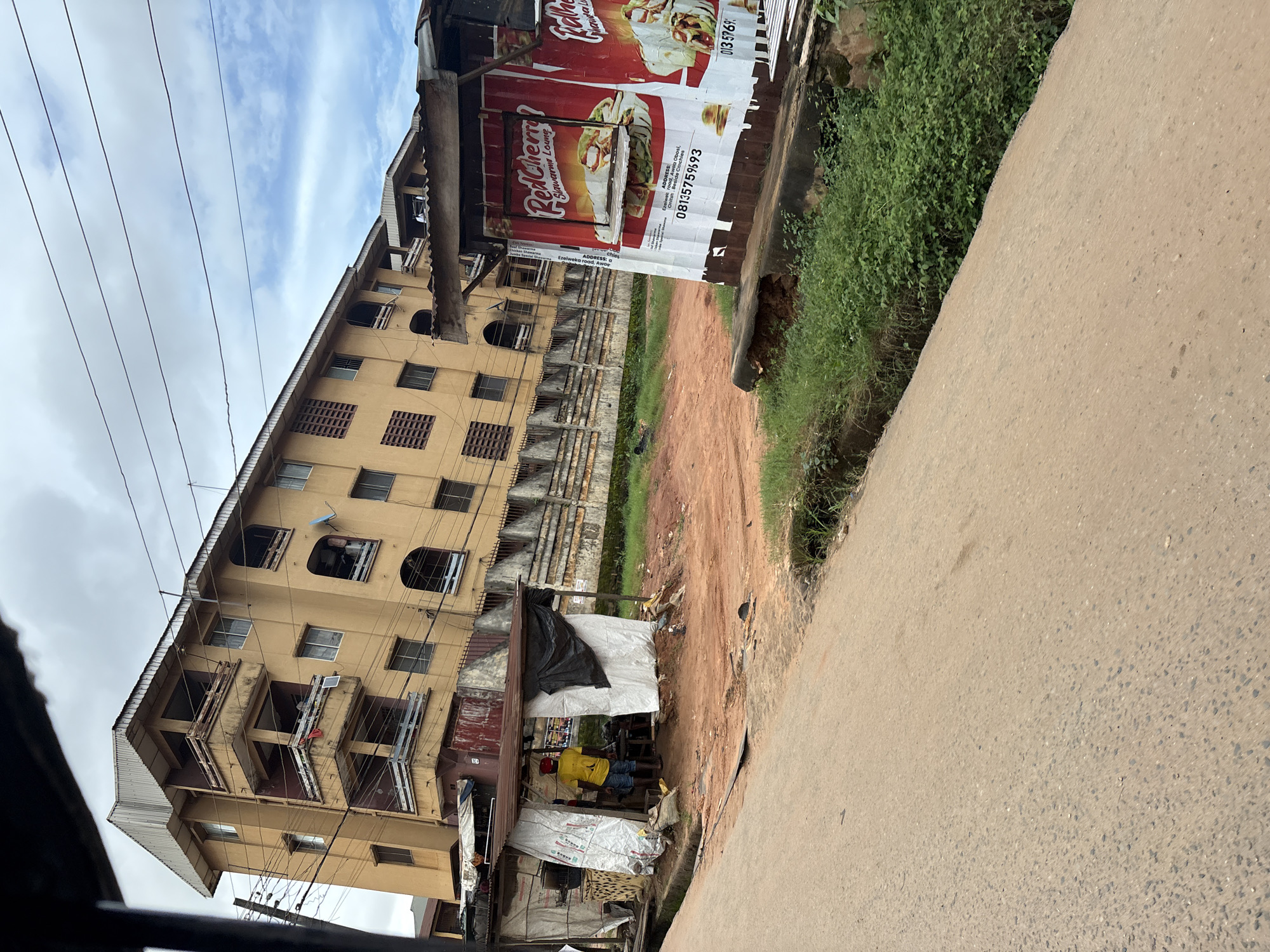 This is a medium-angle, eye-level shot of a street scene in Nkpor, Nigeria, on a cloudy day. In the foreground, a paved road occupies the bottom half of the frame, with its surface showing wear and tear, and a narrow drainage ditch lined with green grass and plants on the right side.
Dominating the middle ground is a reddish-brown, dirt lot. To the left, a makeshift stall is erected, covered with corrugated metal roofing and white tarpaulin sides. Inside, a person wearing a yellow t-shirt and blue shorts stands, facing away from the camera, appearing to be engaged in some activity. To the right of the stall, a tall, narrow container-like structure stands, its exterior covered with large, red advertisements for "Red Cherry Shawarma Lounge," featuring images of shawarma wraps. The phone number "08135759593" is visible on the advertisement.
Behind the lot and stall, a multi-story building stands. It is painted a faded yellow and features multiple balconies with rusty railings. The building has many windows and appears to be residential or commercial. A fence with a decorative pattern runs along the front of the building. Overhead, power lines crisscross the sky, which is filled with gray clouds and patches of blue sky. The overall impression is one of a busy, perhaps developing, urban environment.