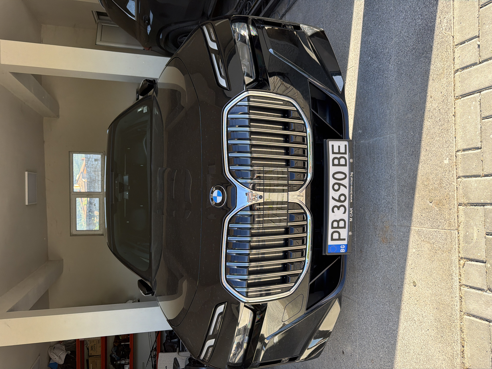The image shows the front of a black BMW car parked indoors, likely a garage, with a Bulgarian license plate reading "PB 3690 BE". The vehicle is located in Asenovgrad, Bulgaria, as suggested by the license plate's "PB" designation and the "www.bmw-mcar.bg" website mentioned on the license plate holder, which is likely a local dealer in Bulgaria. The background shows a garage interior with shelving and various stored items. To the right of the car, there's a paved outdoor area with sunlight.