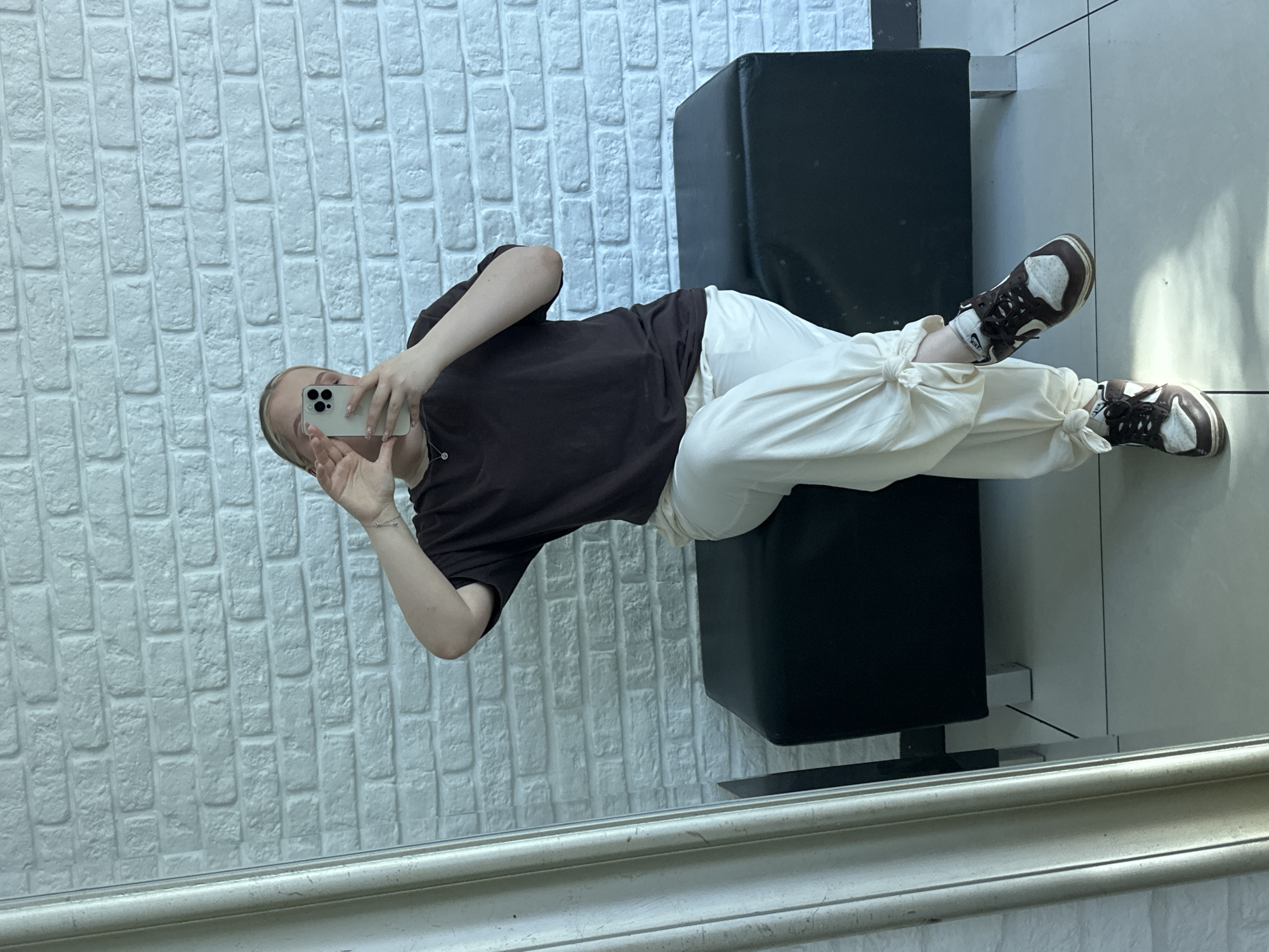 A person is taking a selfie in what appears to be a mirror, positioned at an unusual angle relative to the viewer. The person is wearing a brown short-sleeved shirt and cream-colored pants that are tied at the ankles. On their feet are brown and white sneakers. They are holding a smartphone in their hand, presumably taking the photograph. Behind the person is a wall made of white bricks and a black leather couch or ottoman. The bottom of the mirror and surrounding wall are visible in the bottom of the frame. It is not possible to determine if this photo was taken in the city of Chaikovsky, Russia.