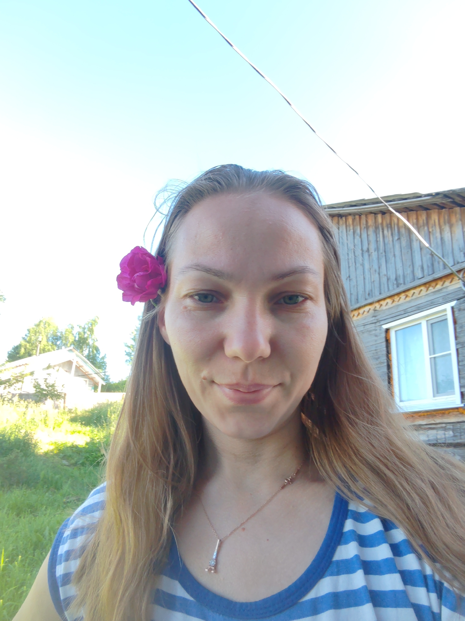 A woman with long blonde hair, adorned with a fuchsia-colored flower behind her right ear, looks directly at the camera. She is wearing a blue and white striped shirt. The background features a wooden building with a white-framed window and a green, grassy area with trees. The sky above is clear and blue. The overall scene suggests an outdoor setting in a rural or suburban area.