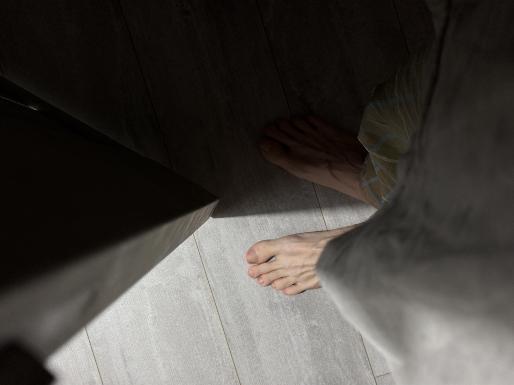 The image shows a close-up, overhead view of two bare feet on a light-colored tiled floor. The feet are positioned on the left and right sides of the frame, with the toes of the left foot clearly visible and the toes of the right foot partially obscured by shadow. The floor appears to be a light gray tile with a wood-grain pattern. A dark, shadowed area, likely a doorway or furniture, is casting a strong shadow across the right side of the floor and partially over the right foot. The lighting suggests an indoor environment, possibly a home or an apartment. There is no information in the image to indicate the city of Naberezhnye Chelny, Russia.